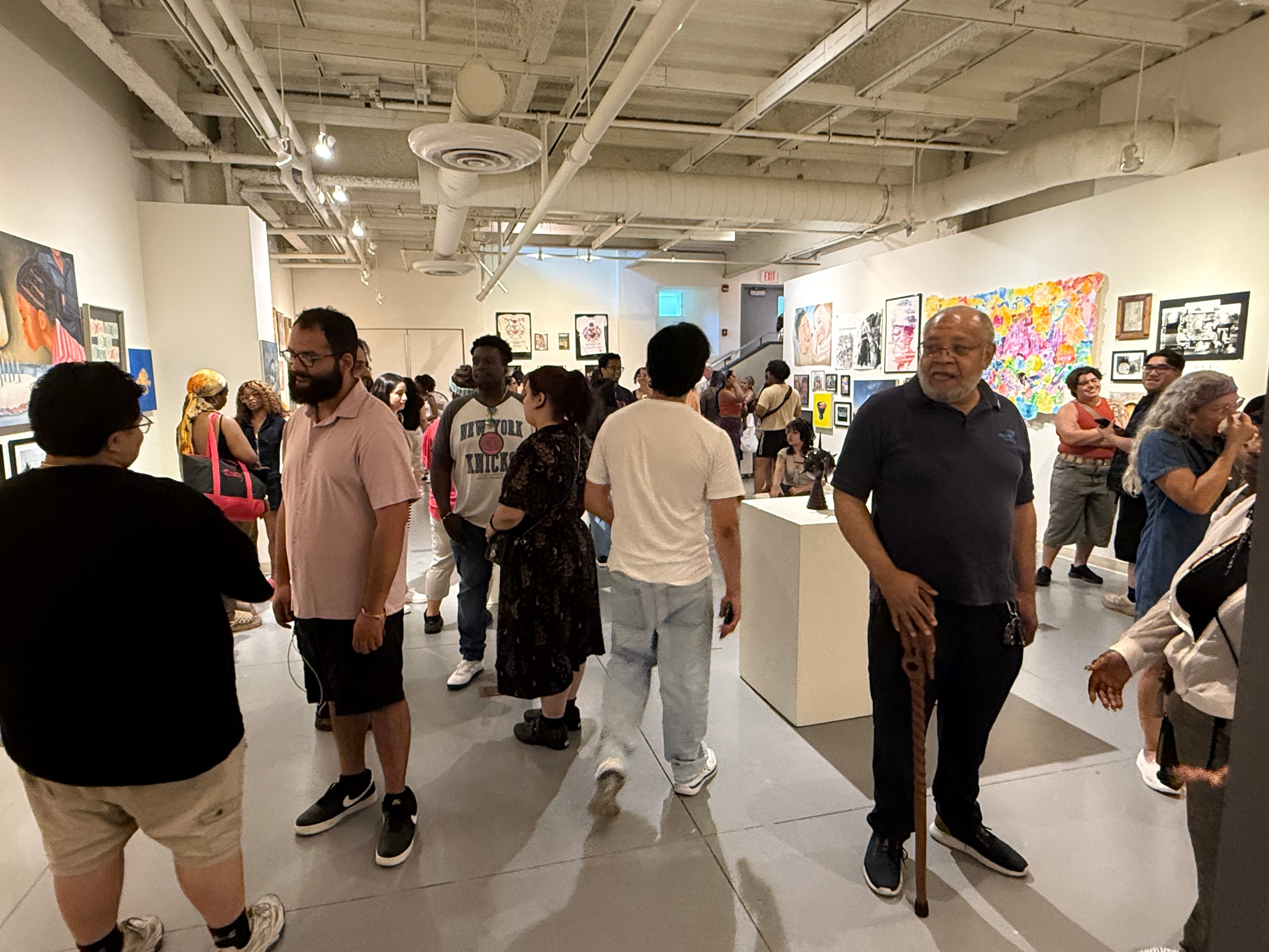 People browsing art gallery with colorful paintings on white walls, diverse group, well-lit modern space, ceiling pipes visible.