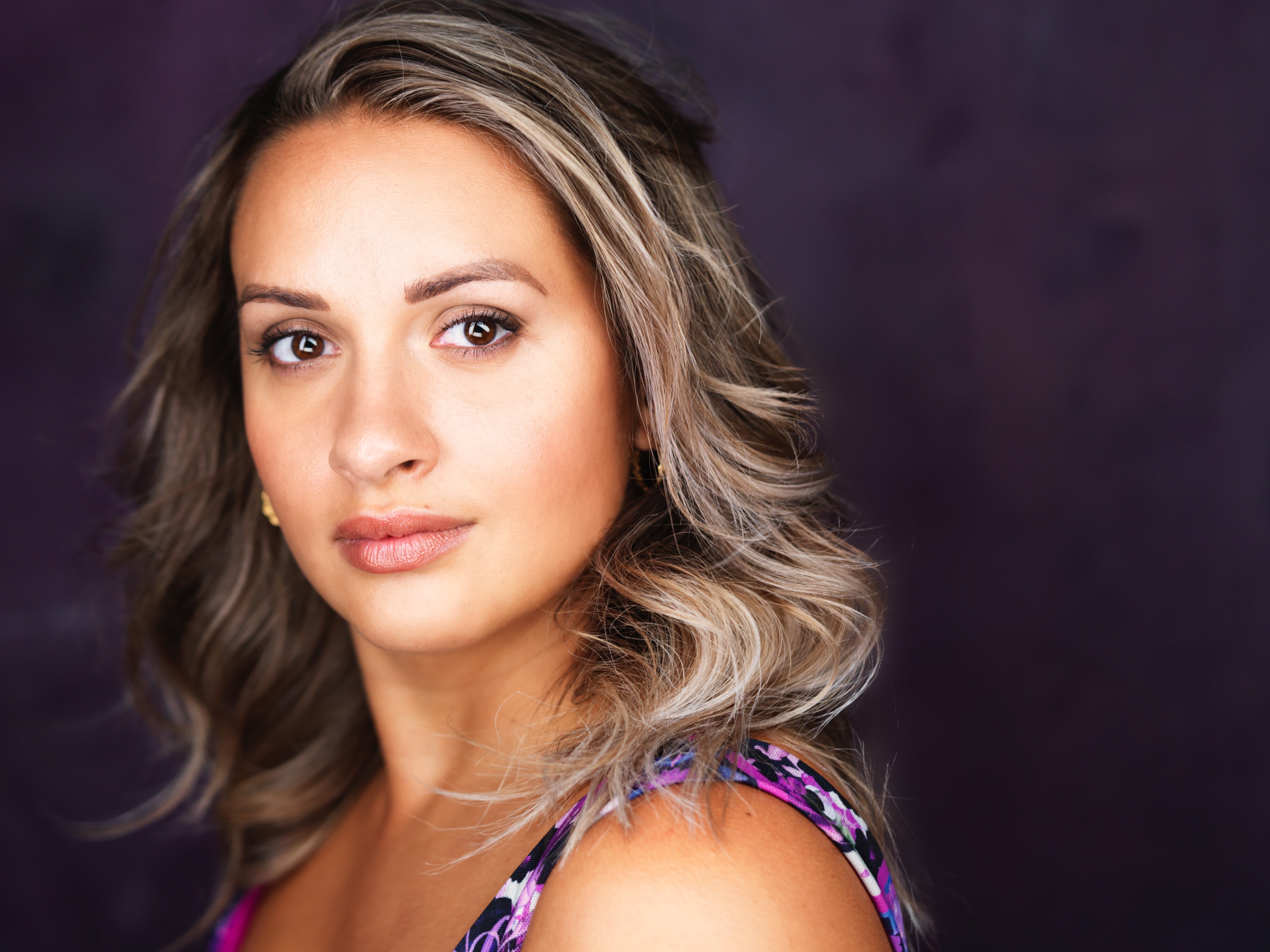 Portrait of a woman with wavy blonde hair, wearing a colorful top, against a dark background.