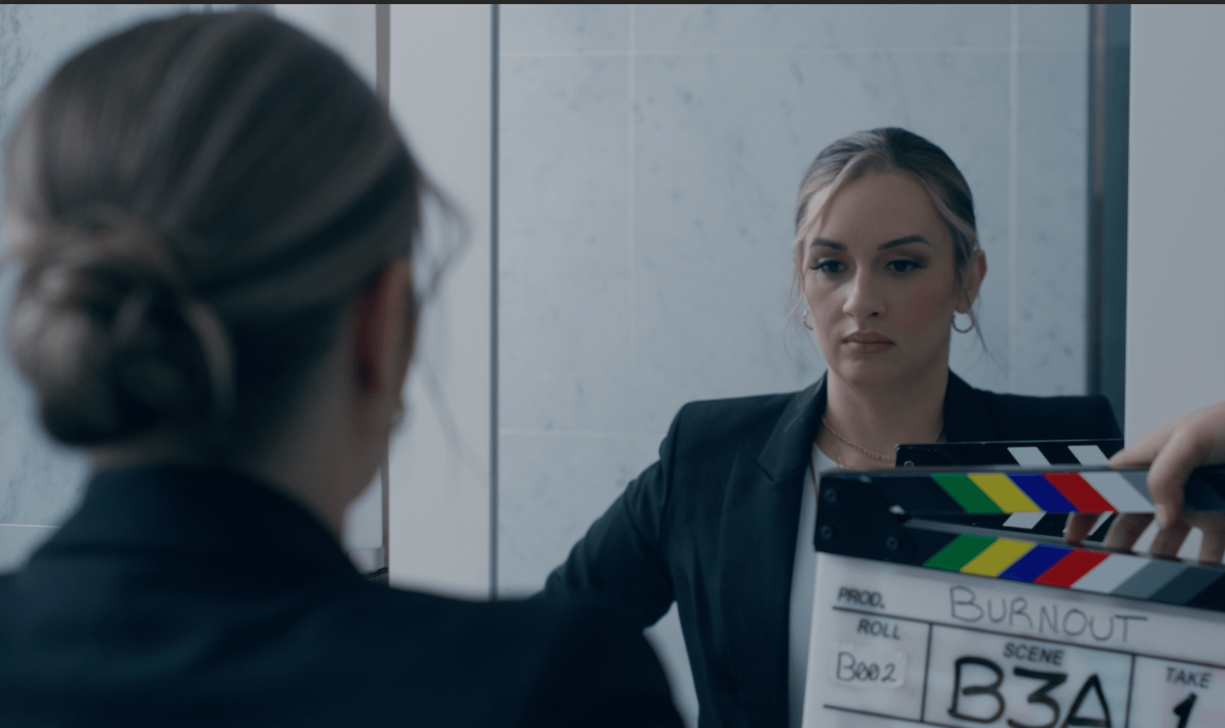 Woman in a dark blazer looks at her reflection in a mirror, holding a clapperboard, in a professional setting.