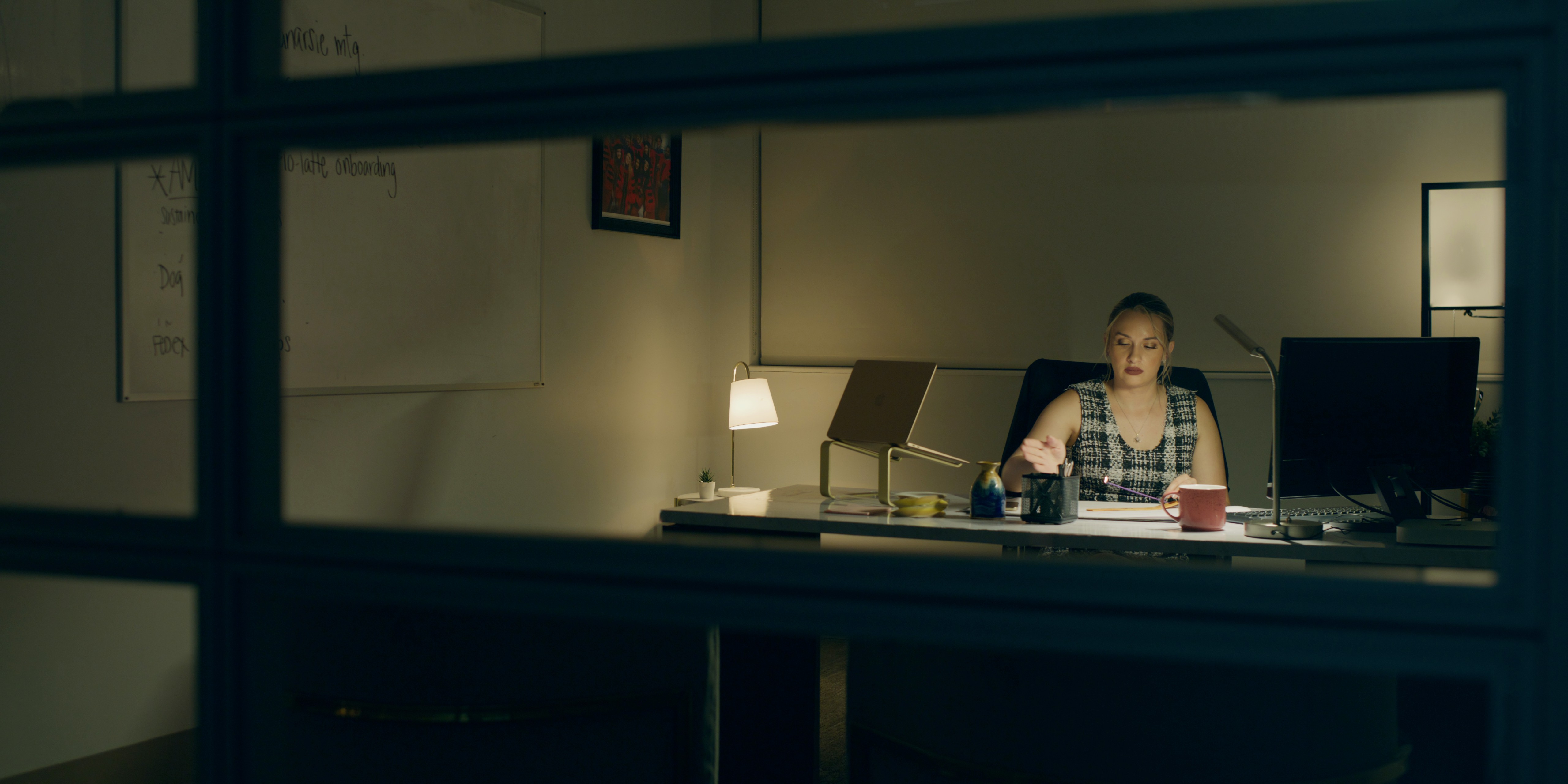 Person sitting at a desk in an office, viewed through a window, with a computer and lamp, in a dimly lit room.
