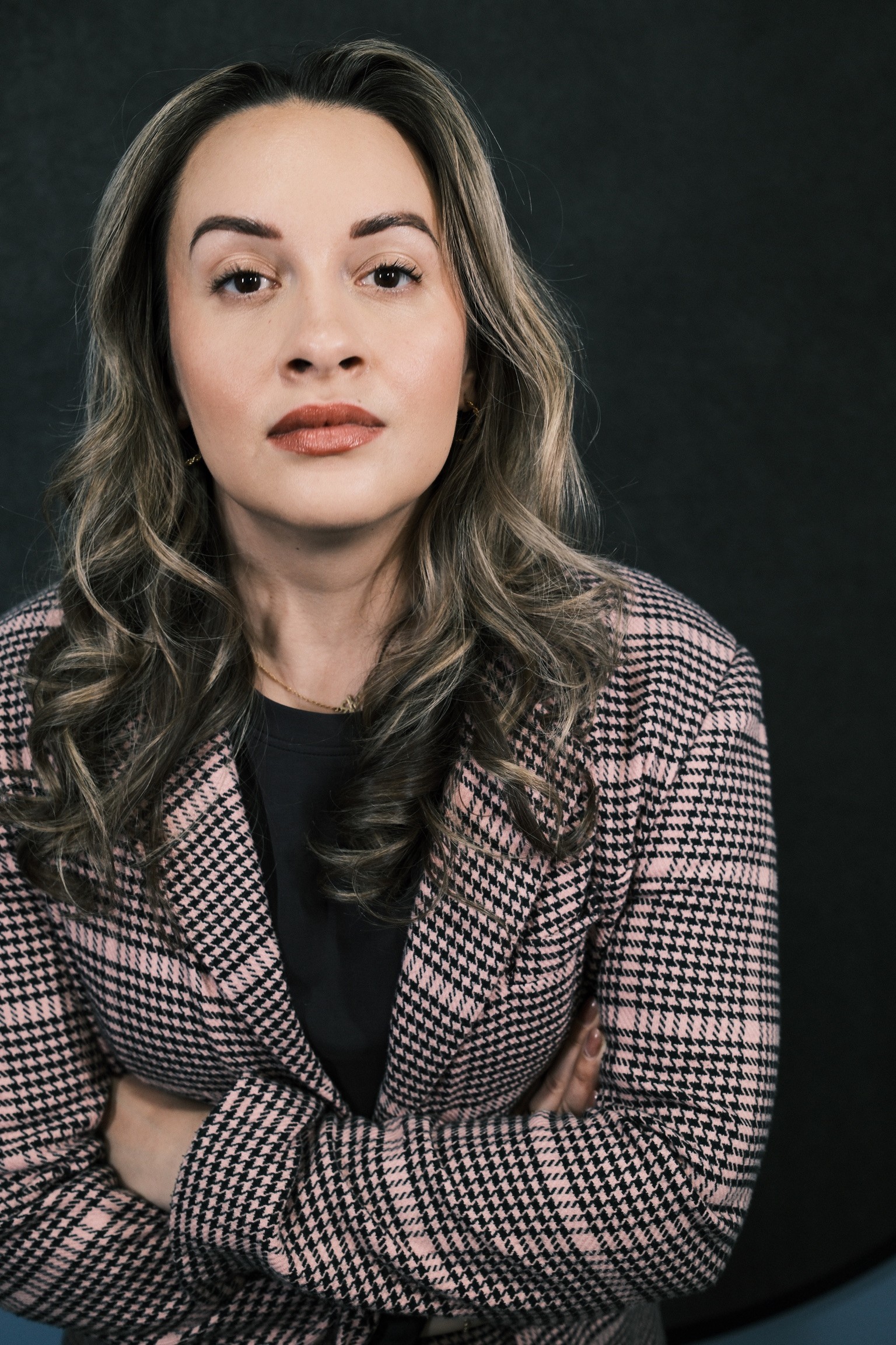 Woman with wavy hair and makeup, wearing a checkered blazer, looking at the camera against a dark background.