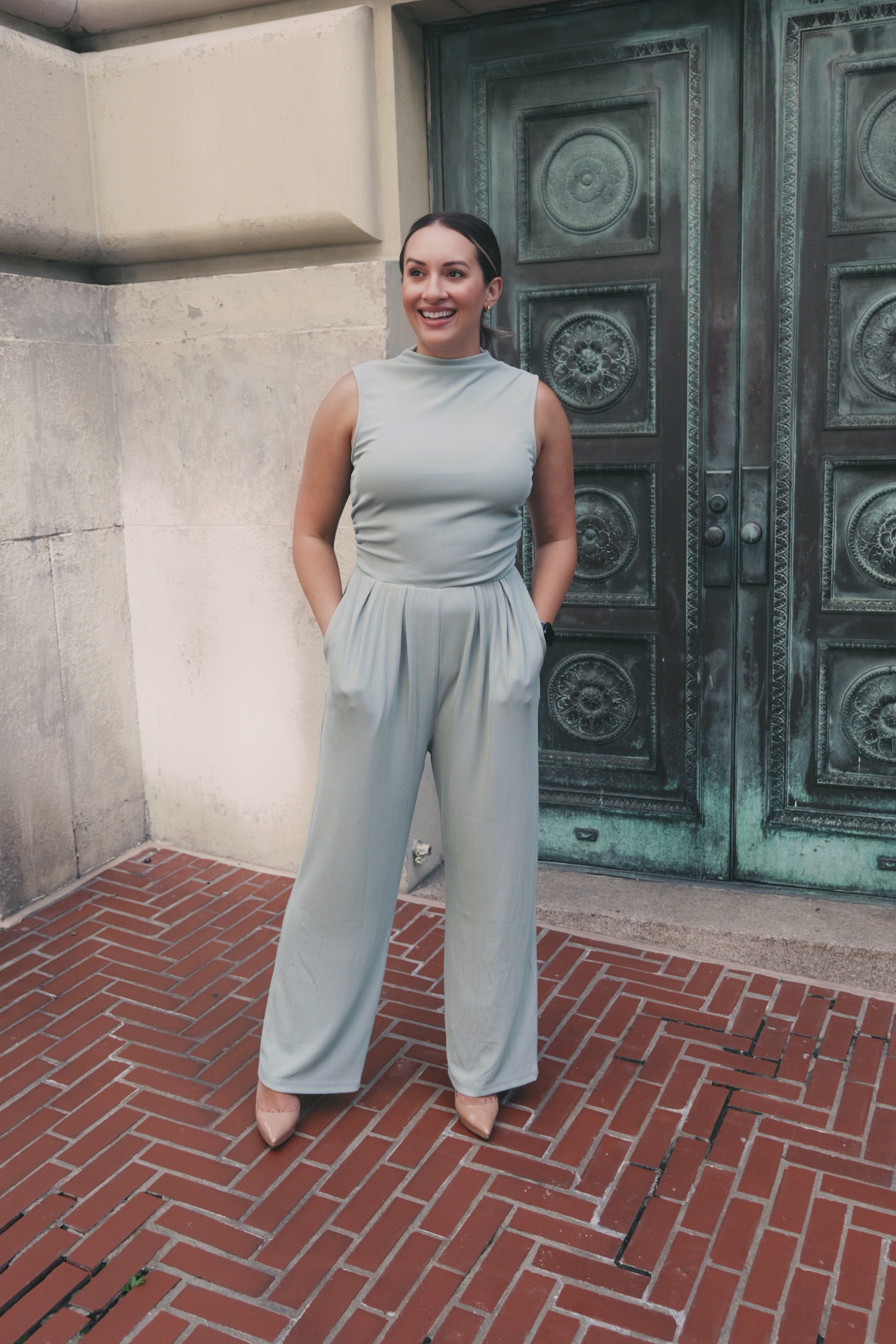 Woman in light-colored sleeveless jumpsuit standing outdoors on brick pavement, smiling, with large ornate door behind her.