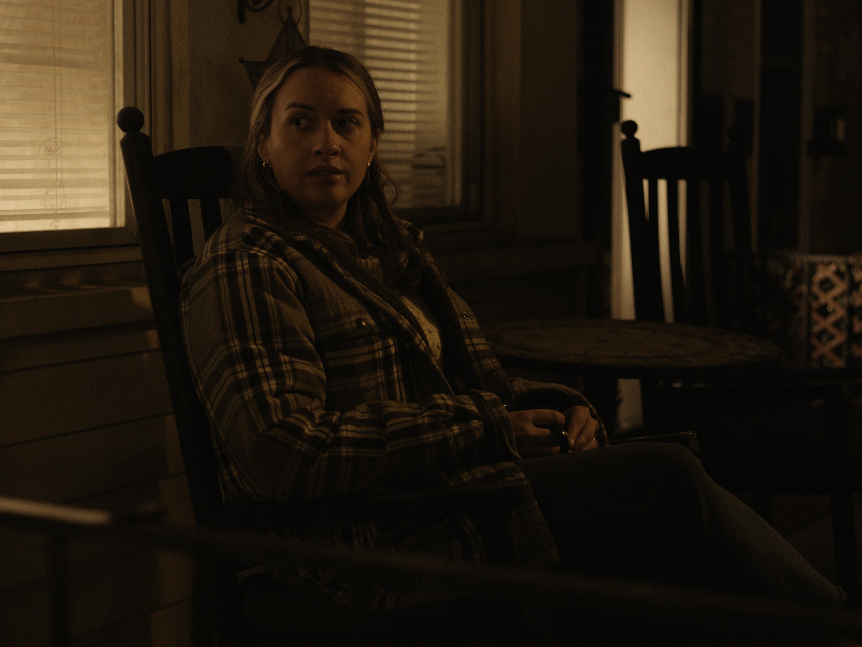 Person sitting in a chair in a dimly lit room with windows and wooden furniture.