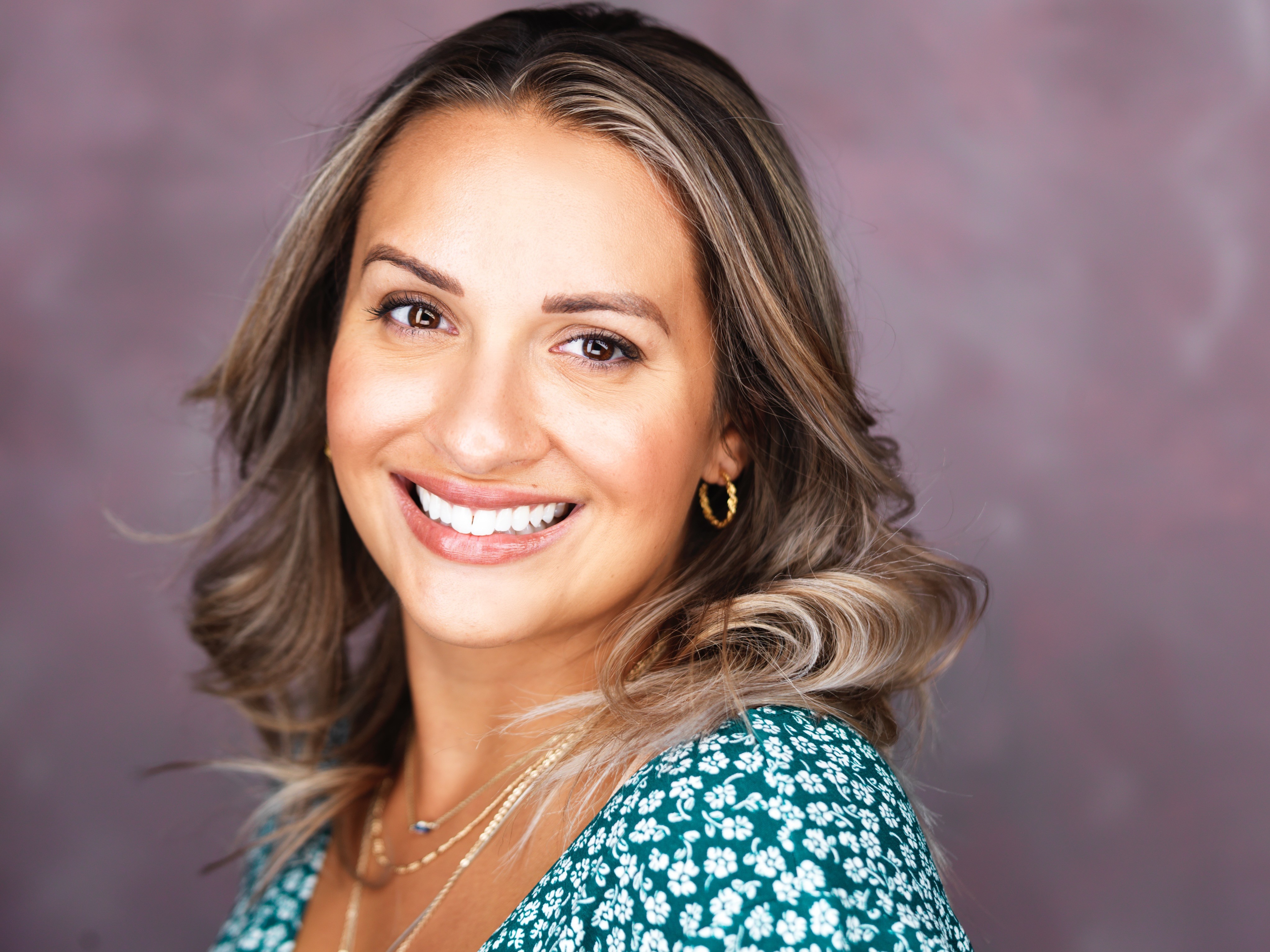 Smiling woman with shoulder-length wavy hair wearing a teal floral top and gold jewelry, against a pinkish background.