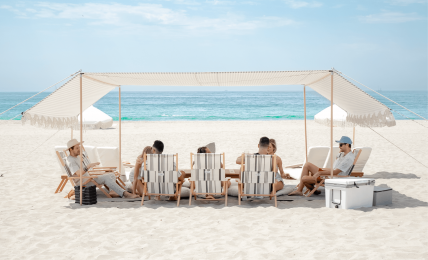 People relaxing under a canopy on a sandy beach with the ocean and sky in the background.