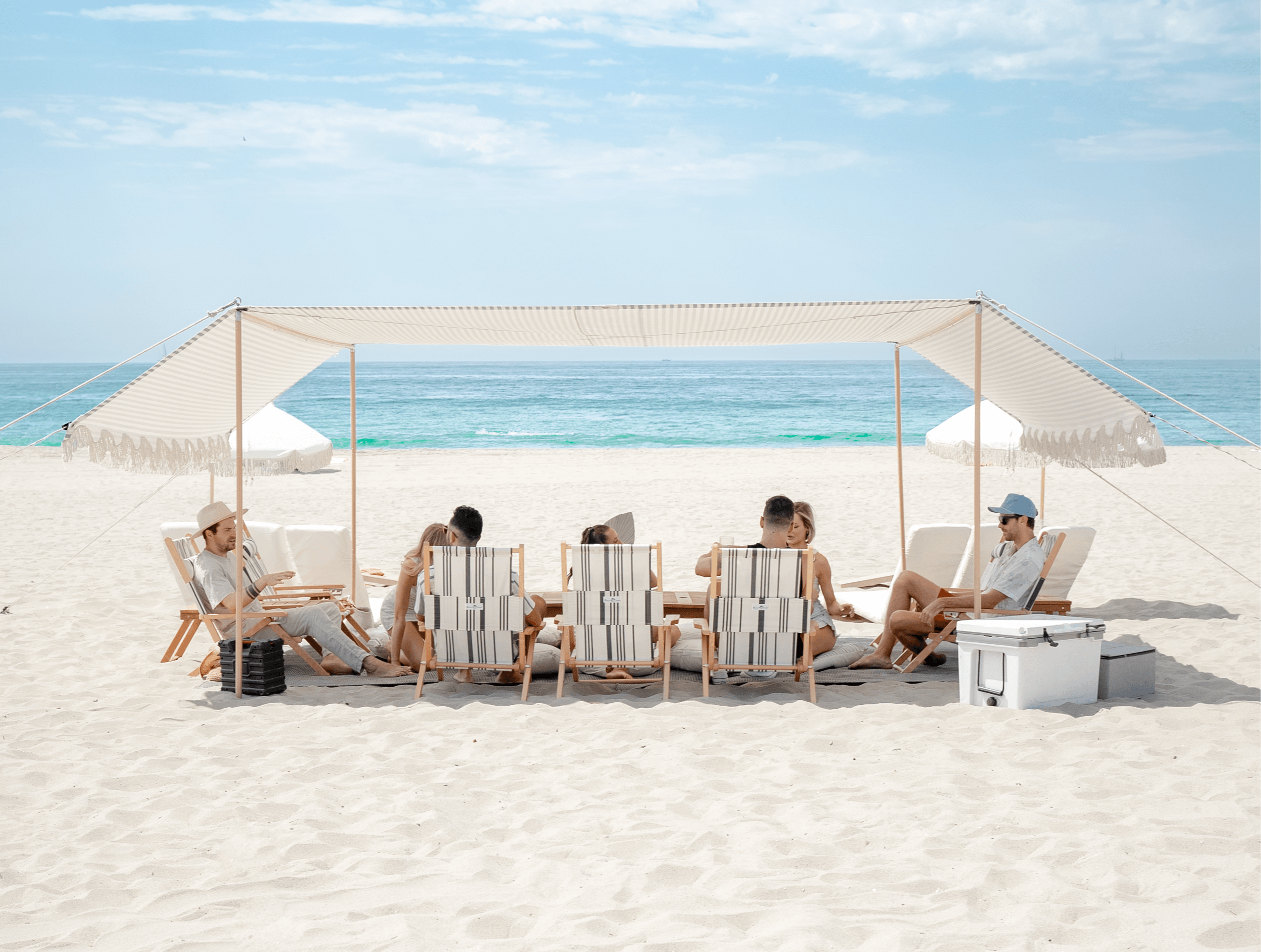 People relaxing under a canopy on a sandy beach with the ocean and sky in the background.