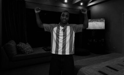 Person standing with arms raised in a room with a sofa and television, black and white image.