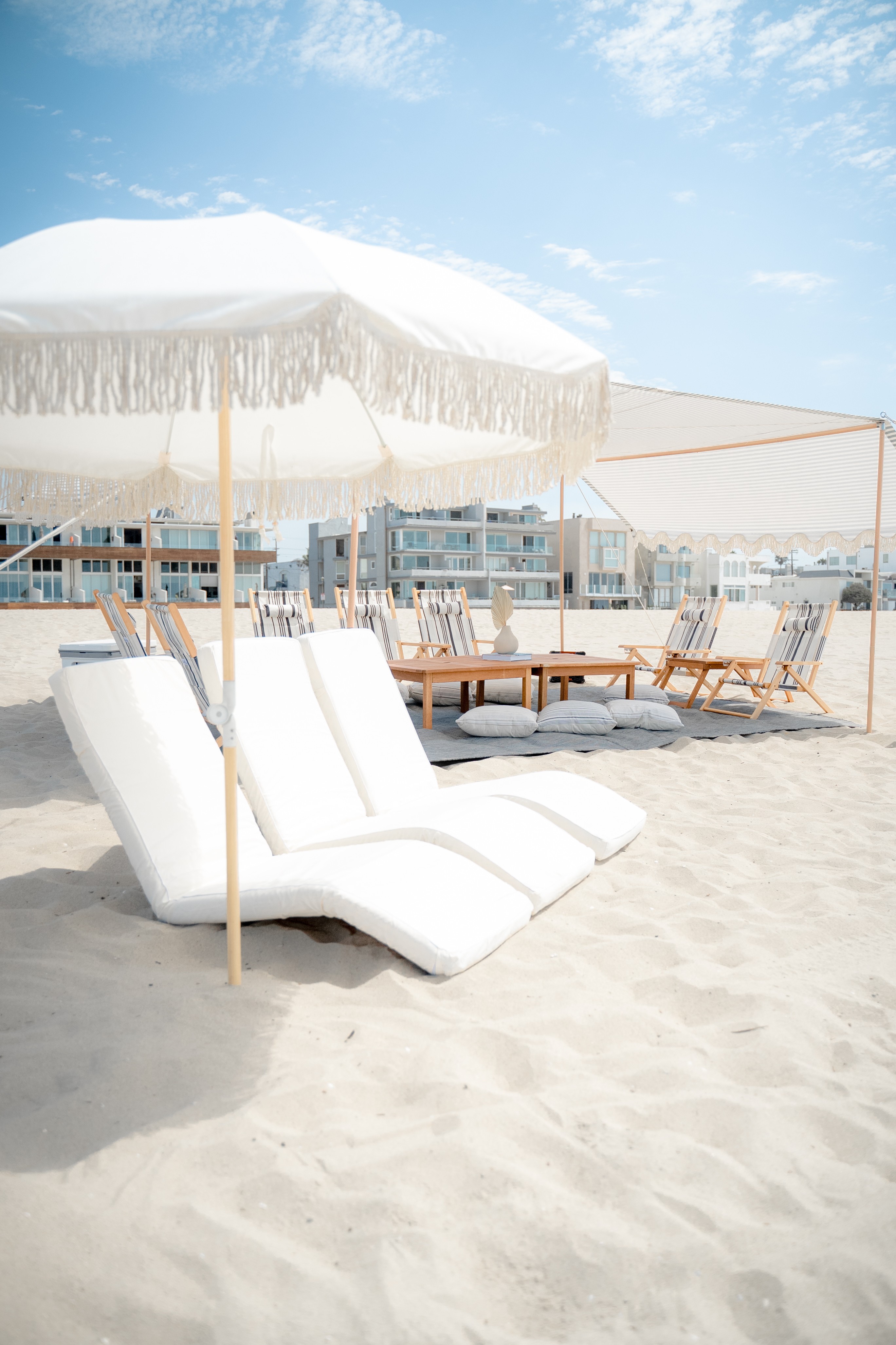 Beach scene with white lounge chairs under a large white umbrella, sandy ground, and a blue sky with clouds.