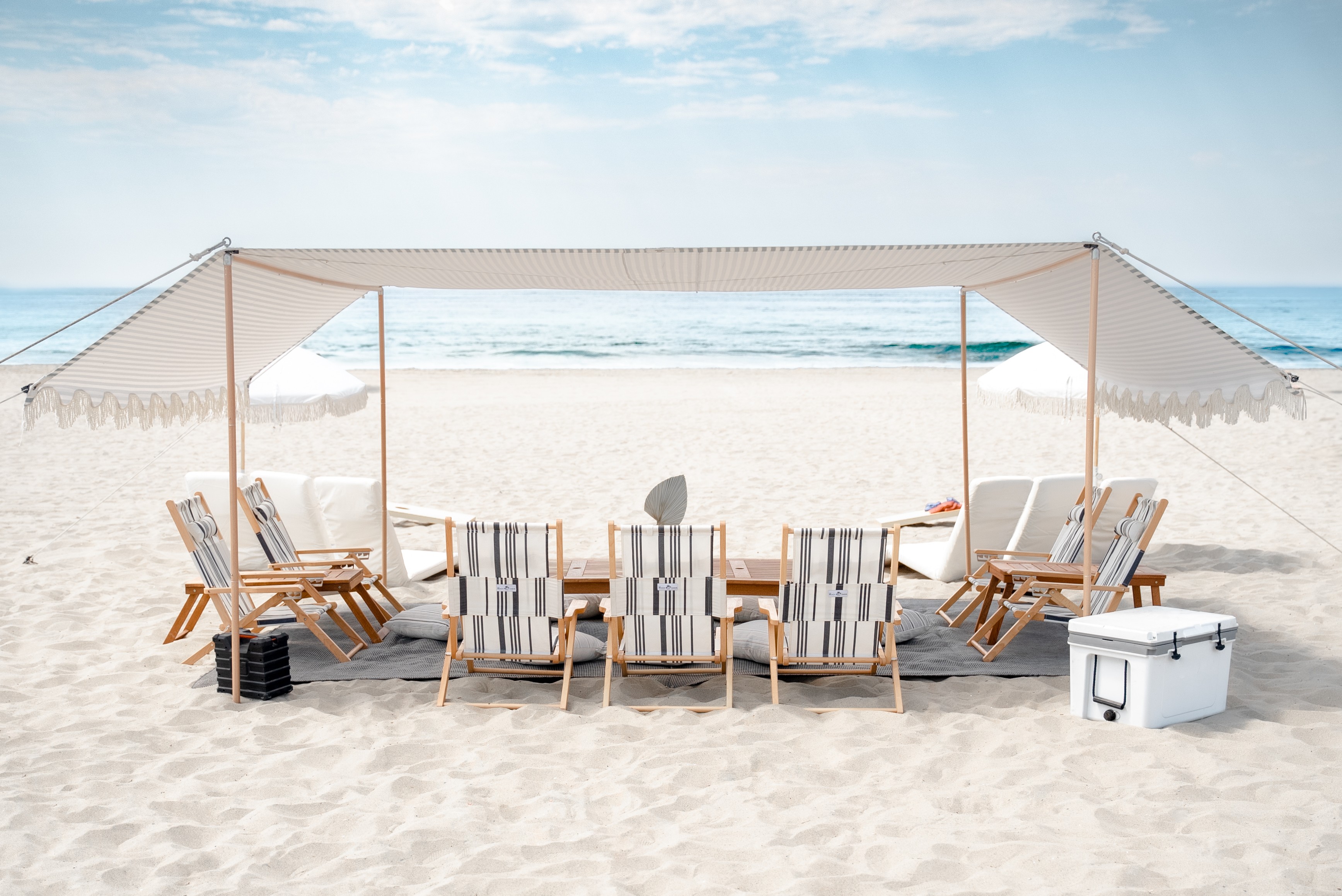 Beach scene with a canopy, wooden chairs, and a cooler on sand, ocean in background, clear sky.