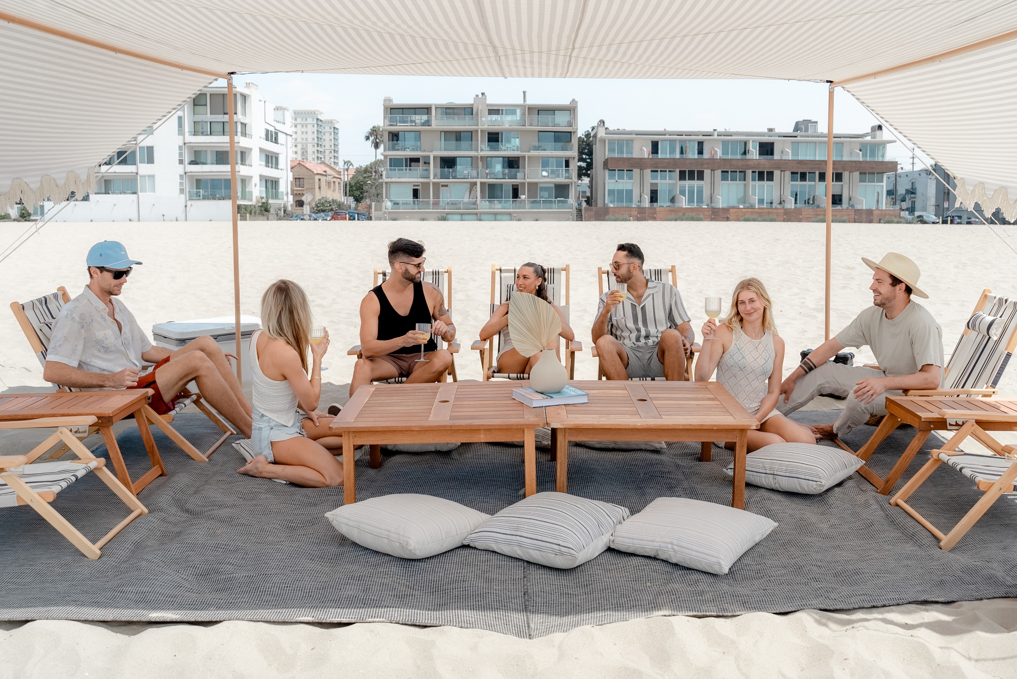 Group of seven people sitting on cushions and chairs under a canopy, with buildings in the background.