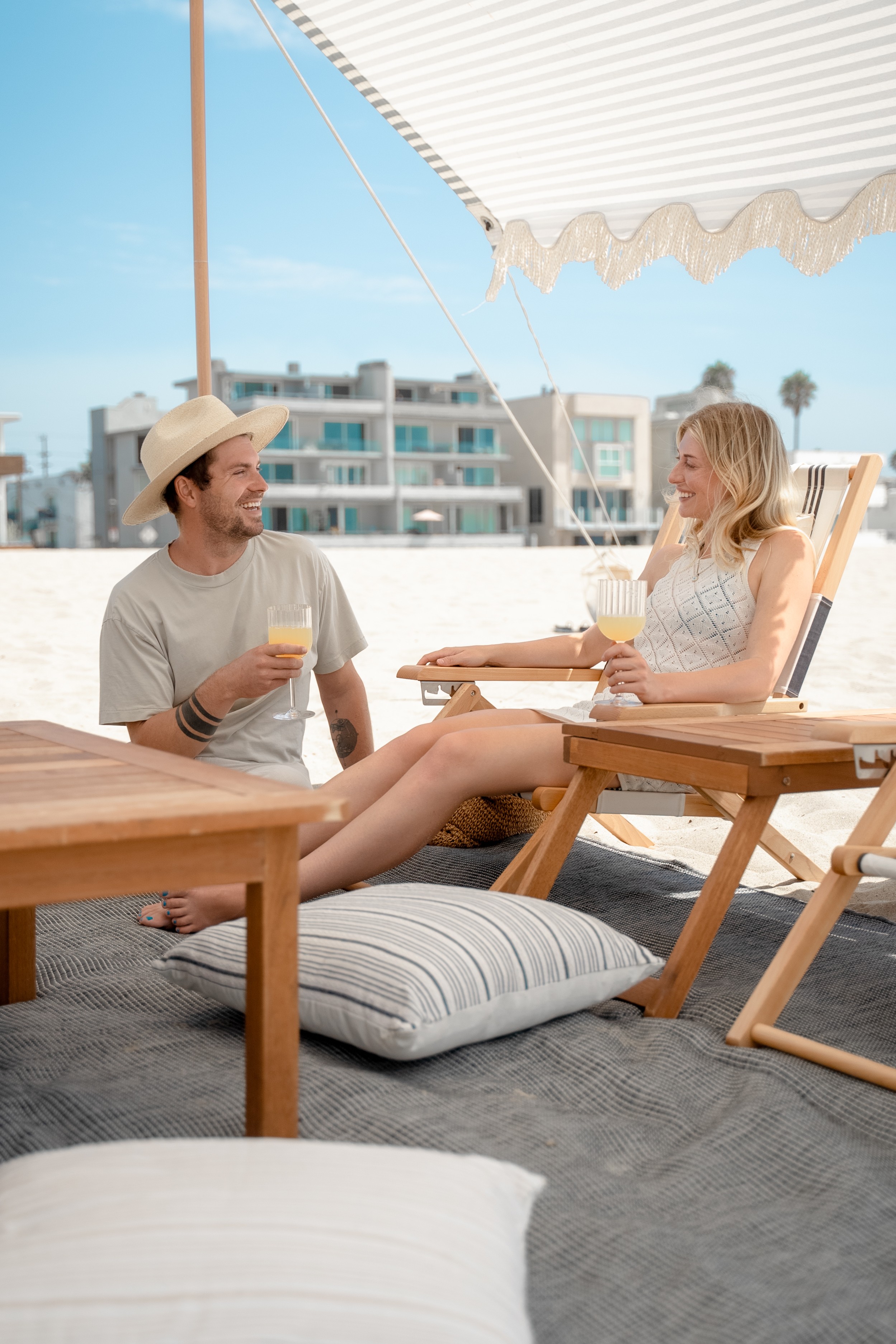 Two people sit on a beach chair under a canopy, enjoying drinks and conversation, with a building and palm trees in background.