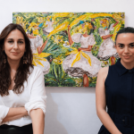 Two women standing in front of a colorful painting, one with long brown hair and white shirt, the other with dark hair and navy dress.