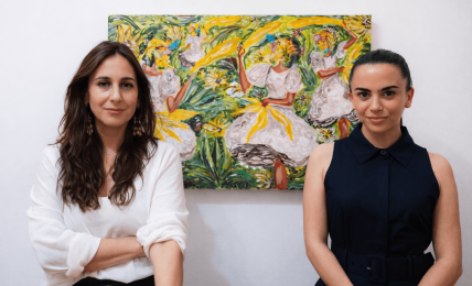 Two women standing in front of a colorful painting, one with long brown hair and white shirt, the other with dark hair and navy dress.