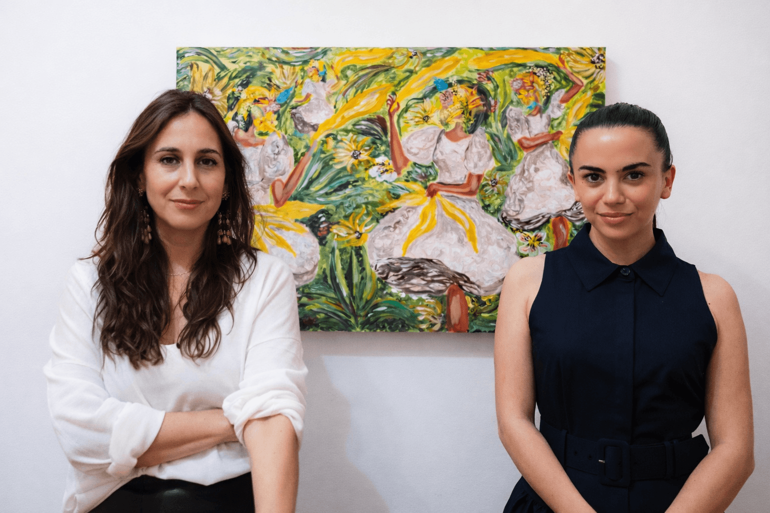 Two women standing in front of a colorful painting, one with long brown hair and white shirt, the other with dark hair and navy dress.