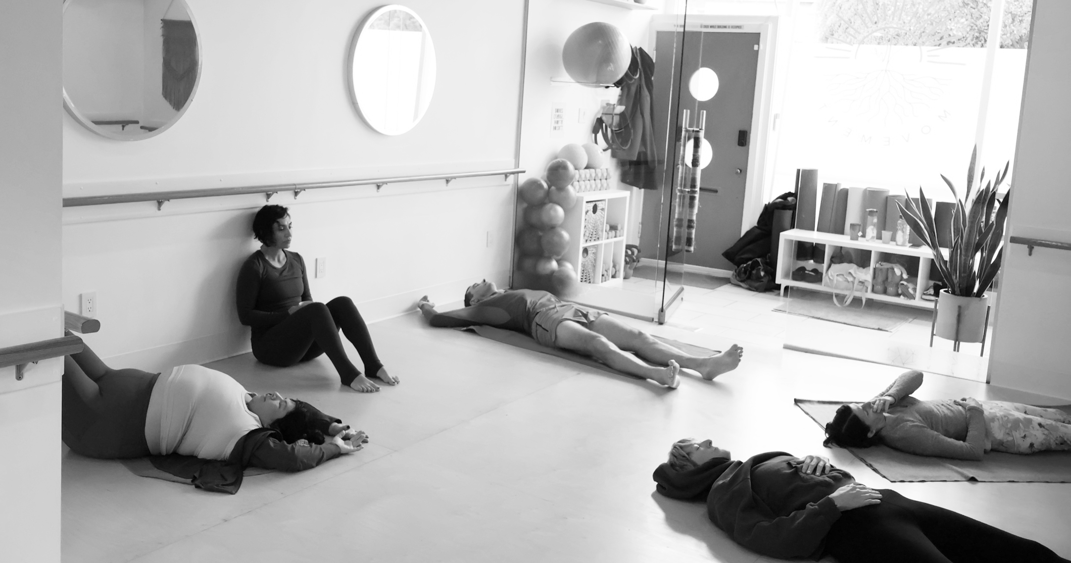 People lying on the floor in a room with mirrors, a person sitting against the wall, and a plant near a window.