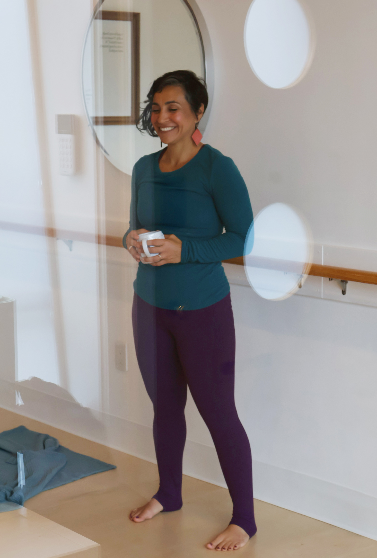 Smiling woman standing barefoot in a room, holding a small object, with a circular window and a handrail behind her.