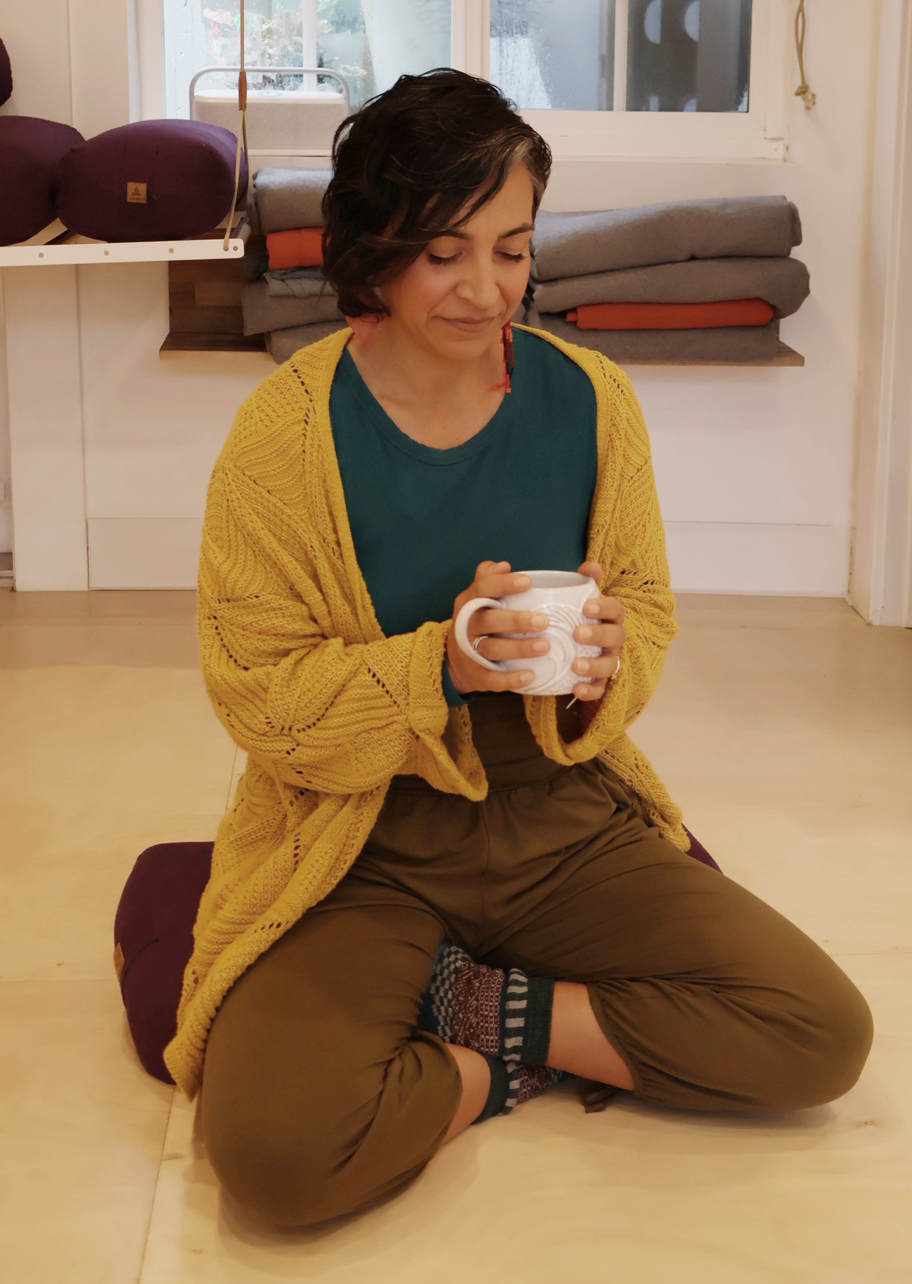 Woman sitting cross-legged on the floor, holding a mug, wearing a yellow cardigan and green top, in a cozy room.