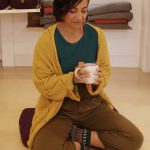 Woman sitting cross-legged on the floor, holding a mug, wearing a yellow cardigan and green top, in a cozy room.