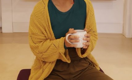 Woman sitting cross-legged on the floor, holding a mug, wearing a yellow cardigan and green top, in a cozy room.