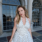 Young woman with wavy hair in a polka dot dress standing outside a building with large arched windows.