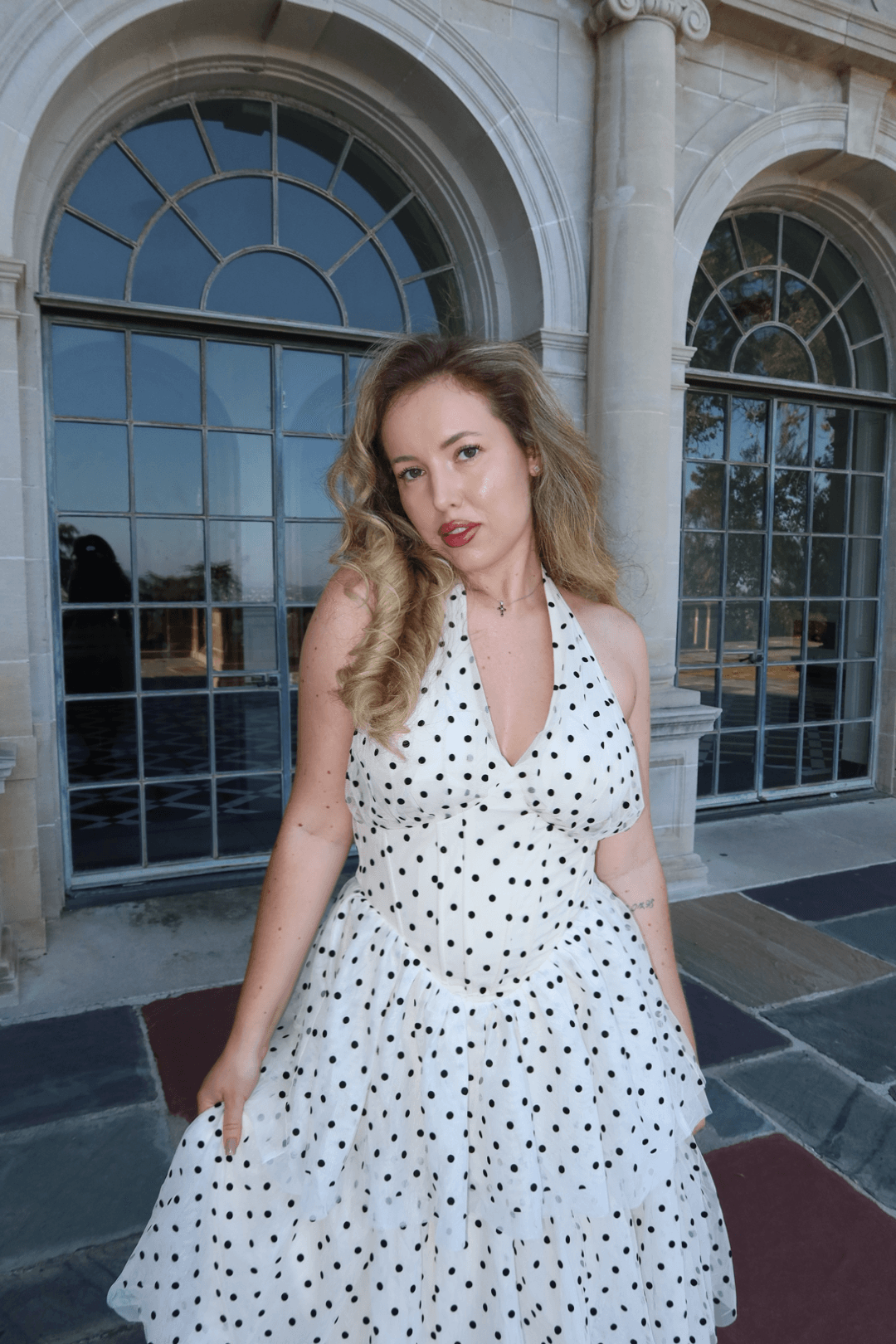 Young woman with wavy hair in a polka dot dress standing outside a building with large arched windows.