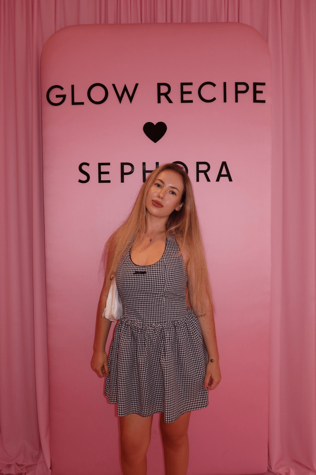 Young woman with long blonde hair standing in front of a pink backdrop with text, wearing a checkered dress.