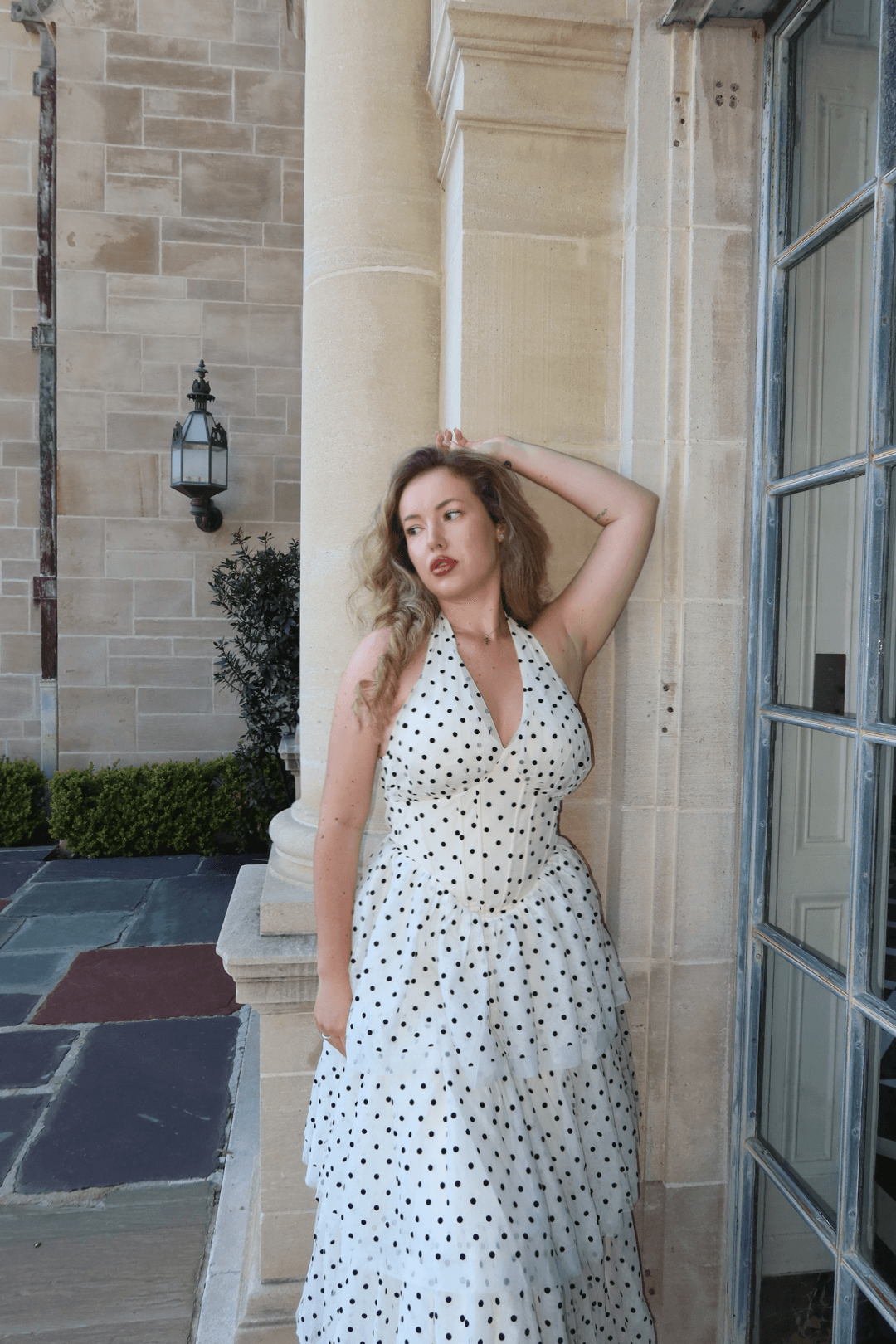 Woman in a white polka dot dress leaning against a stone building with a large window and outdoor lamp, hand on head.