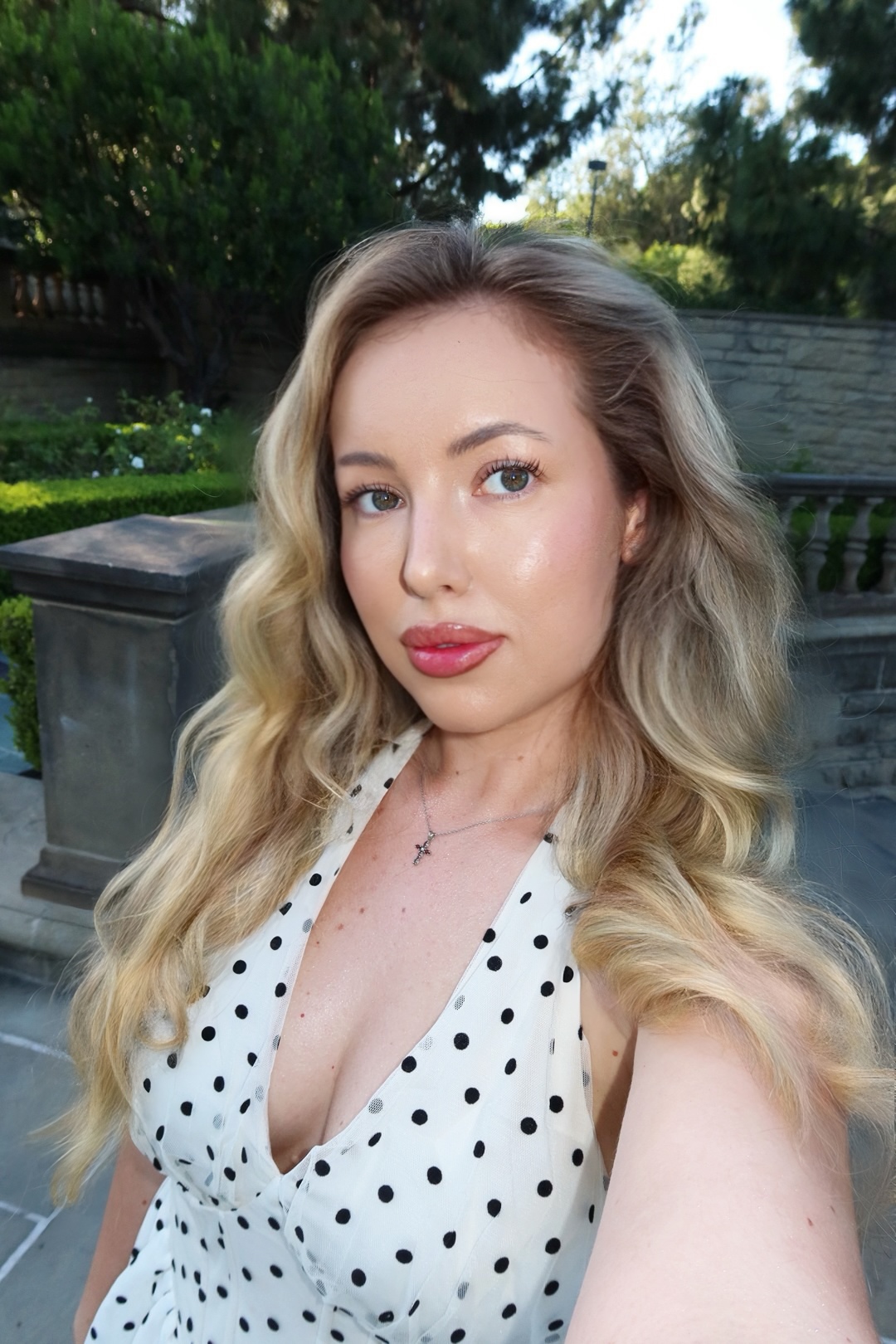 Young woman with wavy blonde hair taking a selfie outdoors, wearing a white polka dot top, with trees and a stone wall in background.