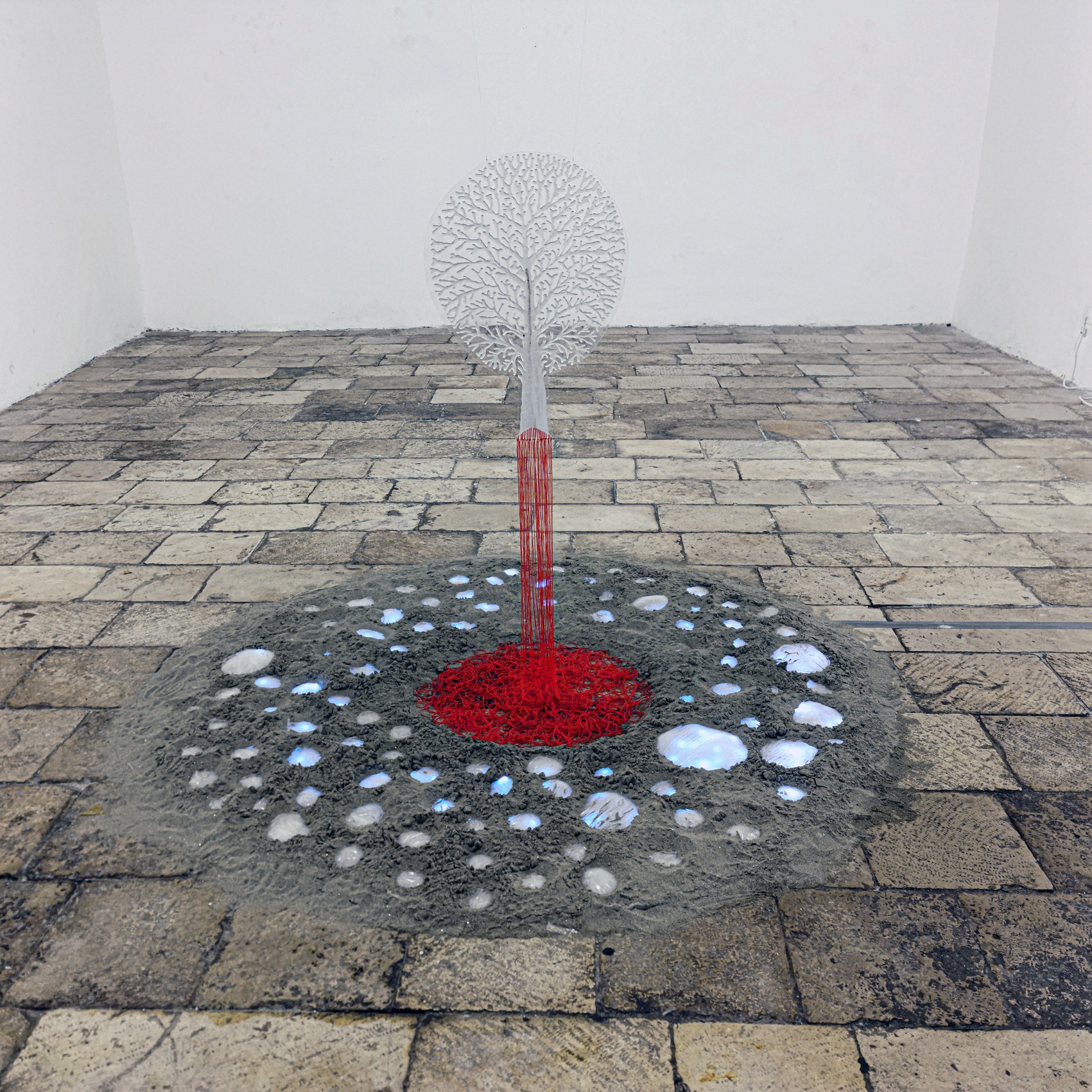 Metal sculpture of a tree with a circular canopy, red trunk, and white base, on a stone-paved floor, against a white wall.