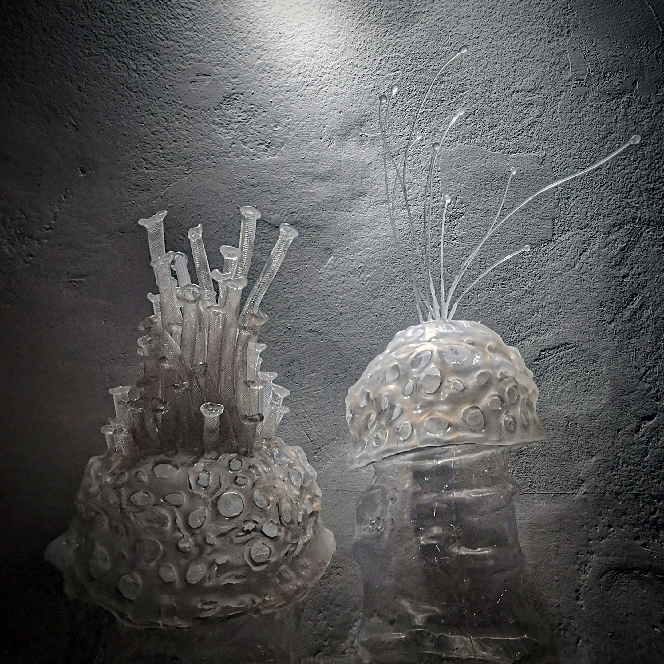 Two glass sculptures with textured bases and delicate, elongated elements extending from the top, set against a dark background.