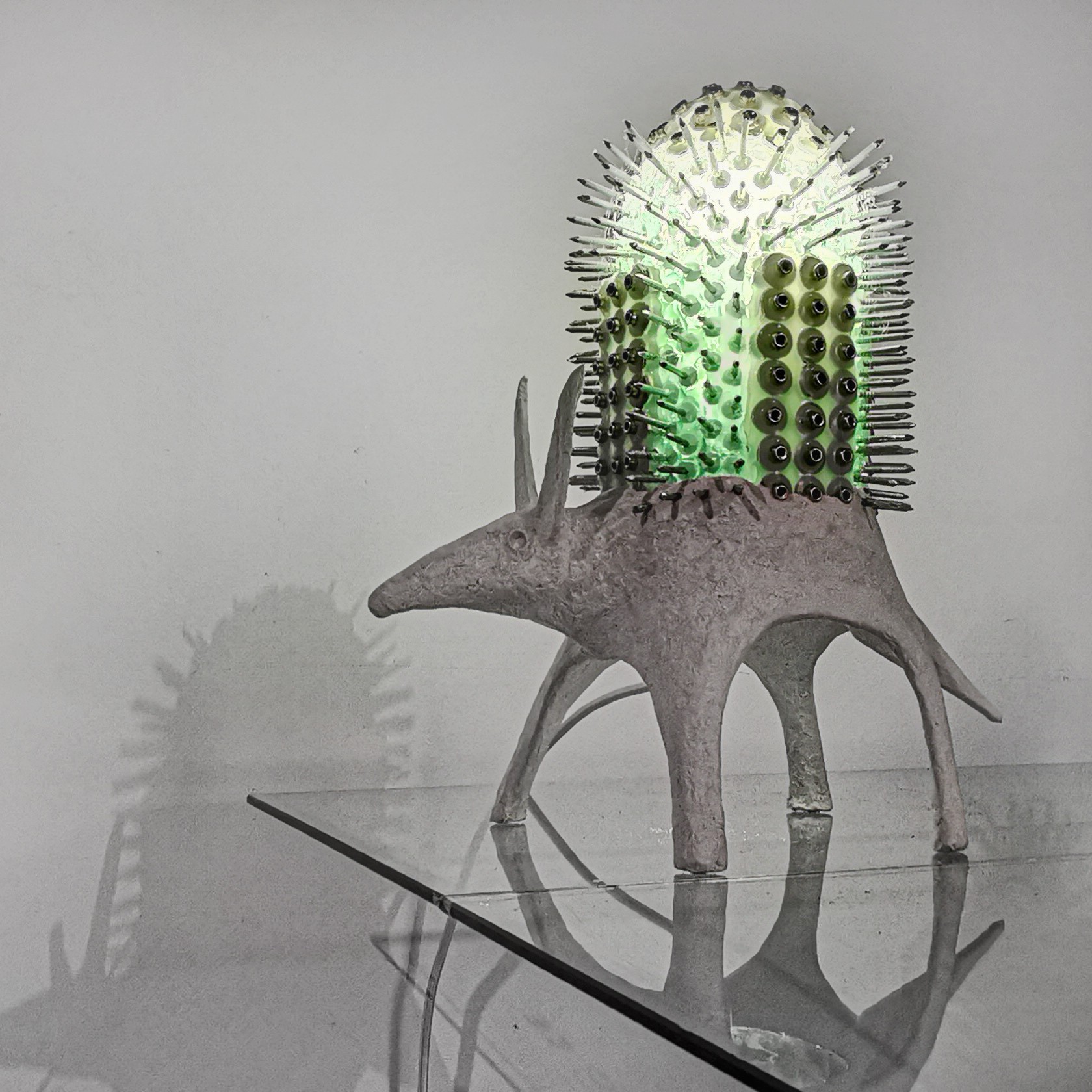 A sculpture of an animal with a cactus on its back, made of various materials, standing on a reflective surface.