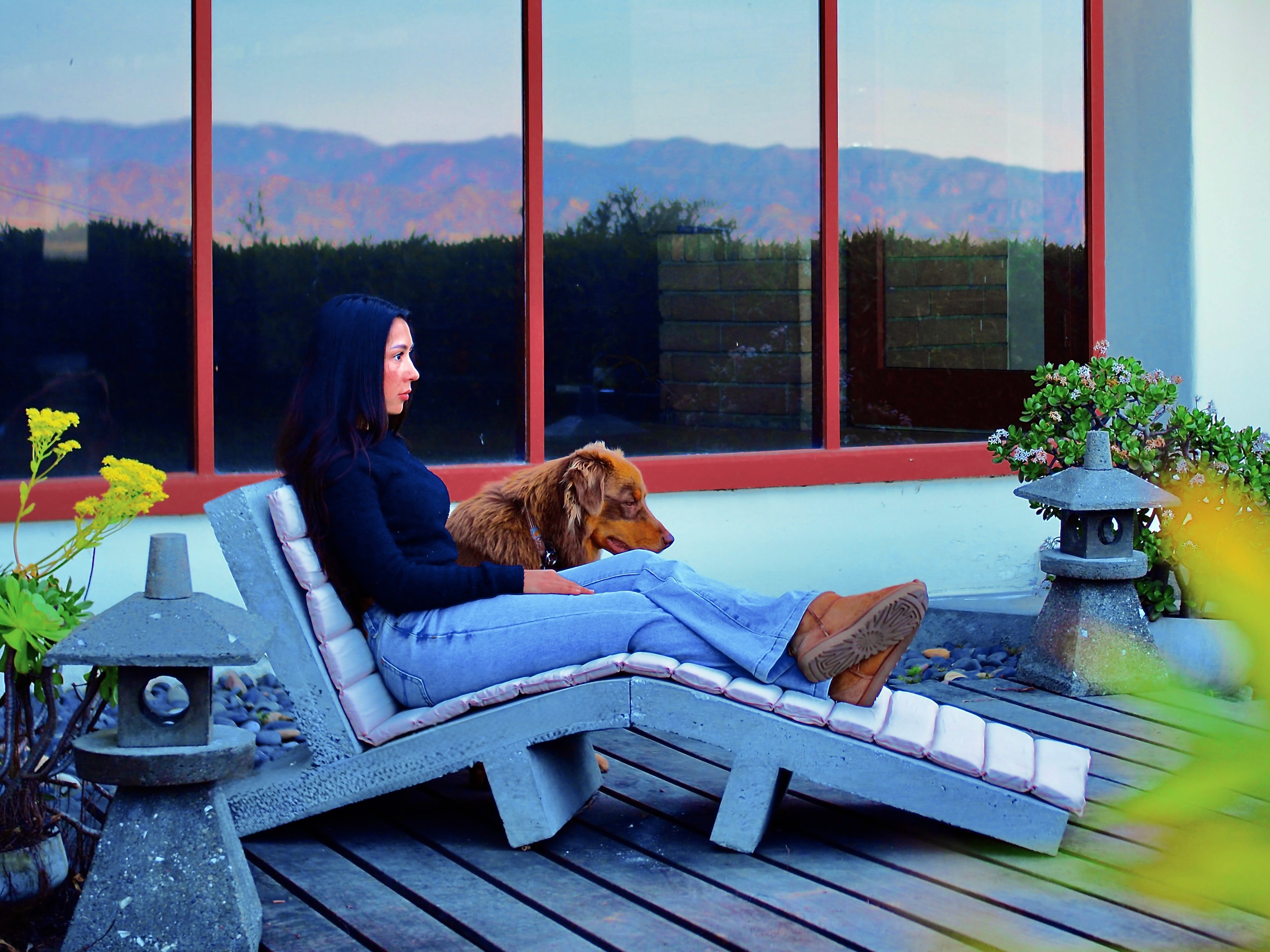 Woman with a dog relaxing on a lounge chair outdoors, with large windows and mountains in the background.