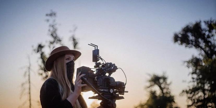 Person wearing a hat and mask operating a camera outdoors during sunset or sunrise.