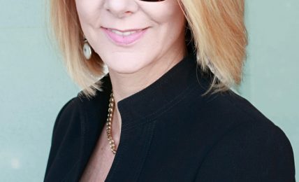 Woman with blonde hair and glasses smiling, wearing a black blazer, against a light background.
