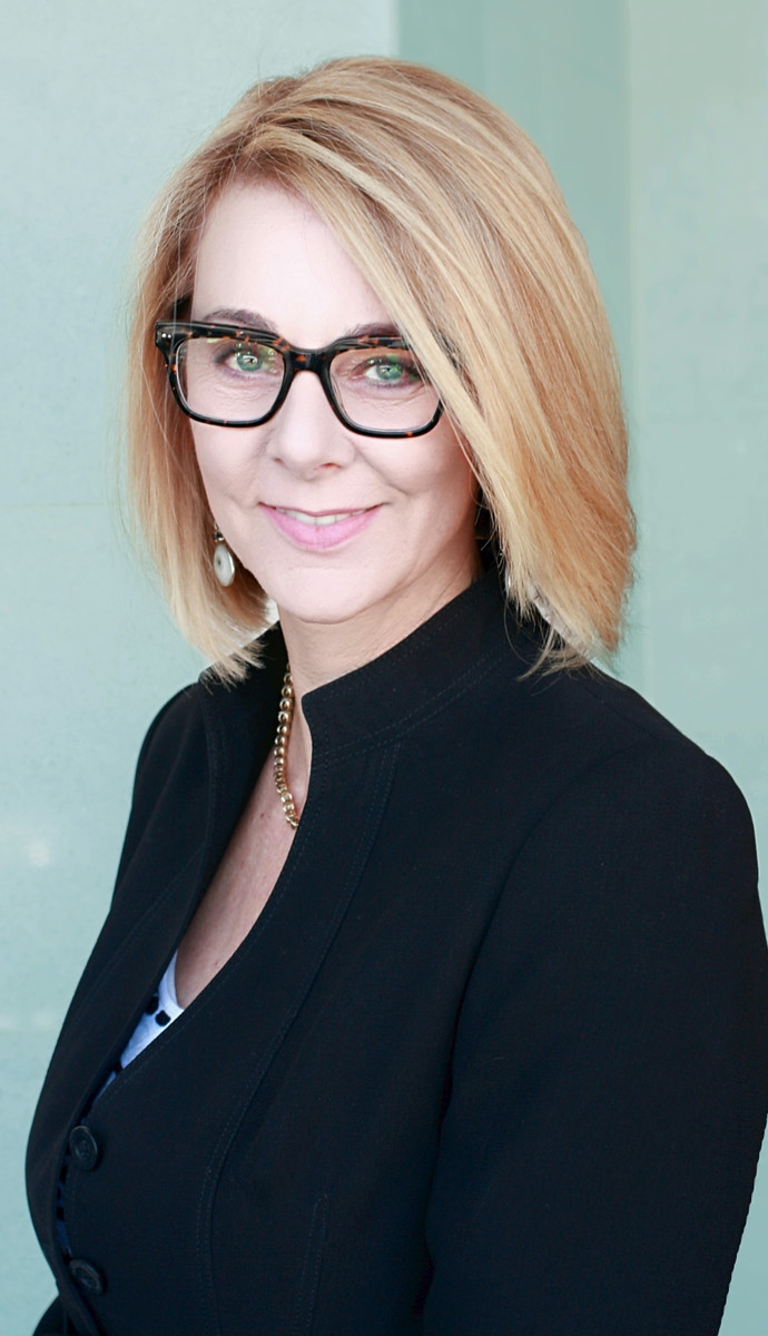 Woman with blonde hair and glasses smiling, wearing a black blazer, against a light background.
