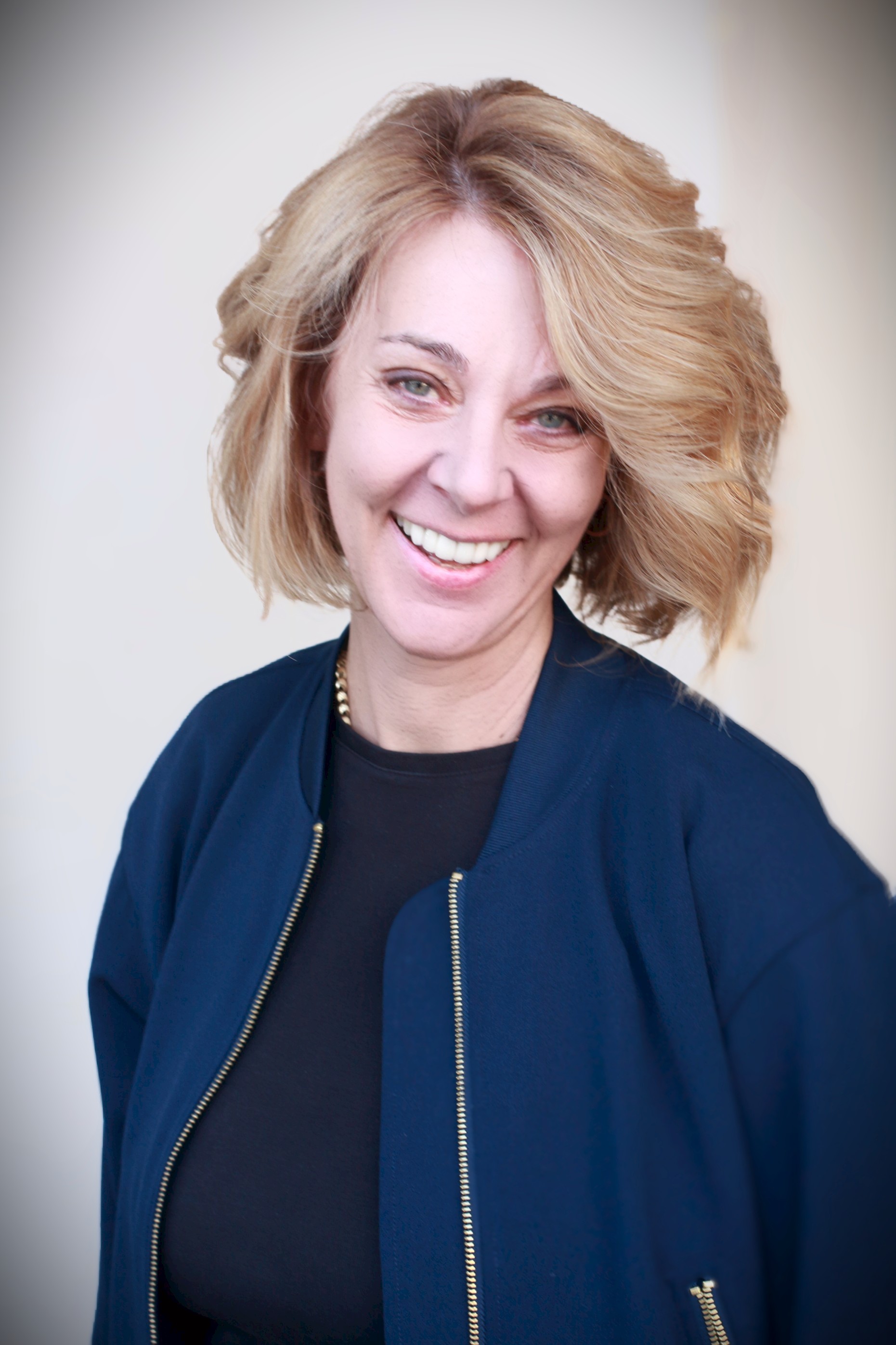 Woman with blonde, wavy hair smiling, wearing a dark top and a navy blue jacket, against a light background.