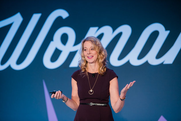 Woman with blonde hair smiling, holding a microphone and a remote, standing in front of a large 'Woman' sign.