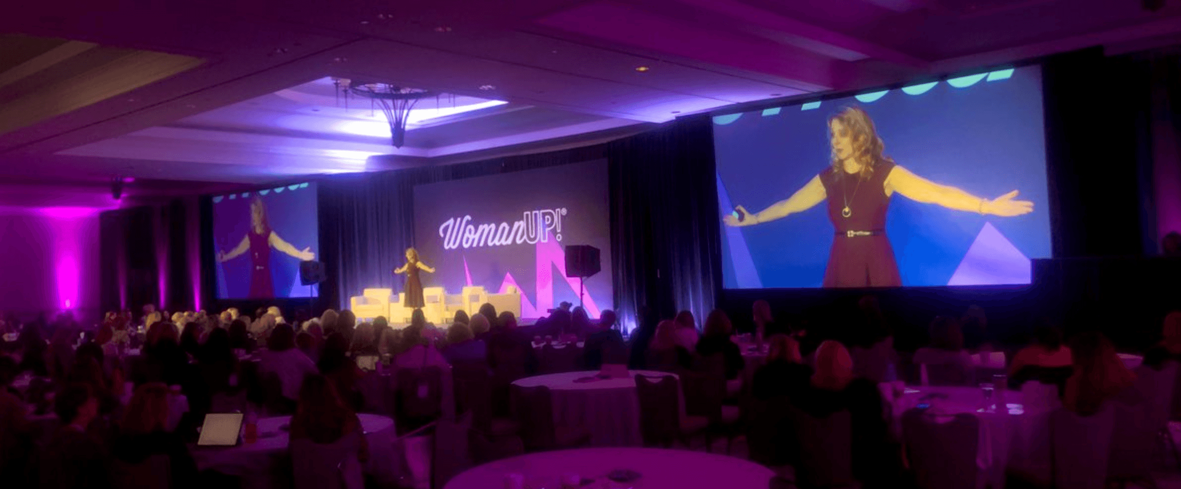 Conference room with a stage, large screens showing a woman speaking, audience seated, purple lighting, and a sign that says 'Welcome'.