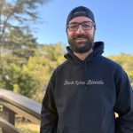 Smiling man with glasses, beard, black hoodie, and backwards cap outdoors with trees and blue sky.