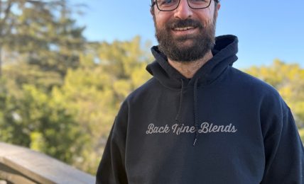 Smiling man with glasses, beard, black hoodie, and backwards cap outdoors with trees and blue sky.