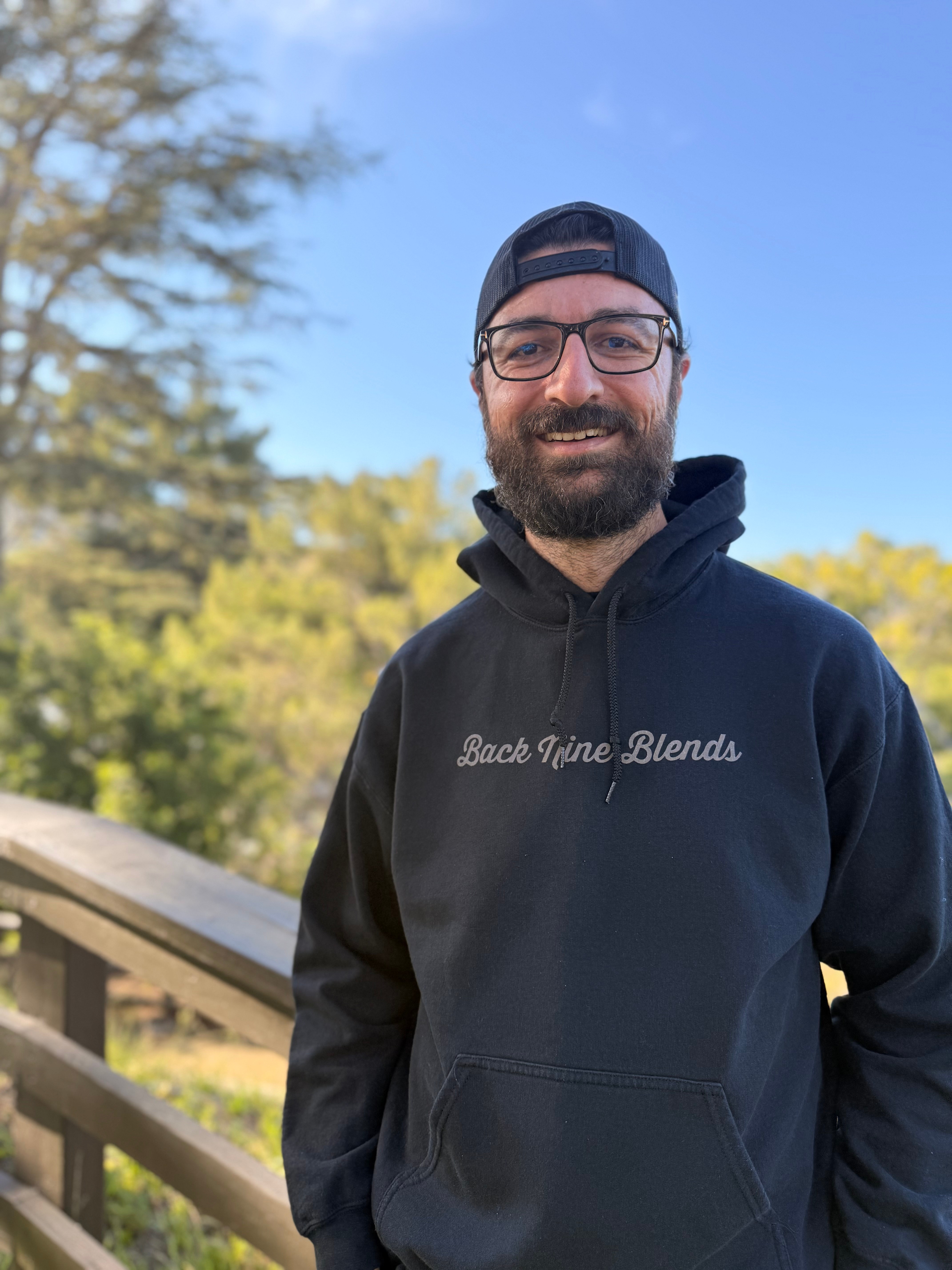 Smiling man with glasses, beard, black hoodie, and backwards cap outdoors with trees and blue sky.