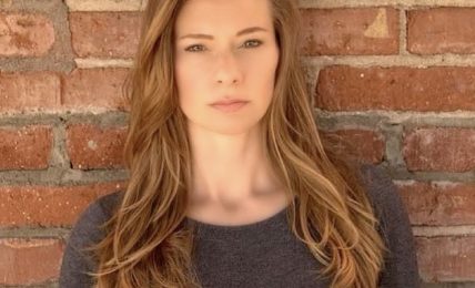Young woman with long, wavy hair standing against a brick wall, wearing a dark gray top.