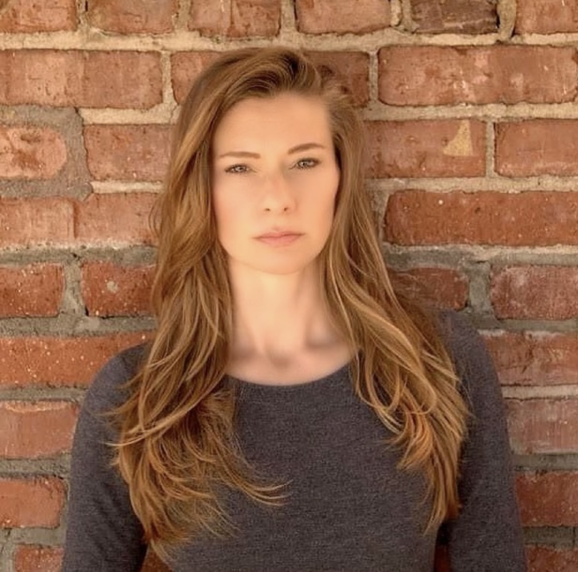 Young woman with long, wavy hair standing against a brick wall, wearing a dark gray top.