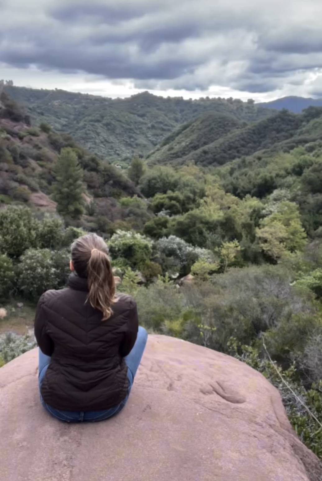 Person sitting on a rock overlooking a green valley with hills and cloudy sky.