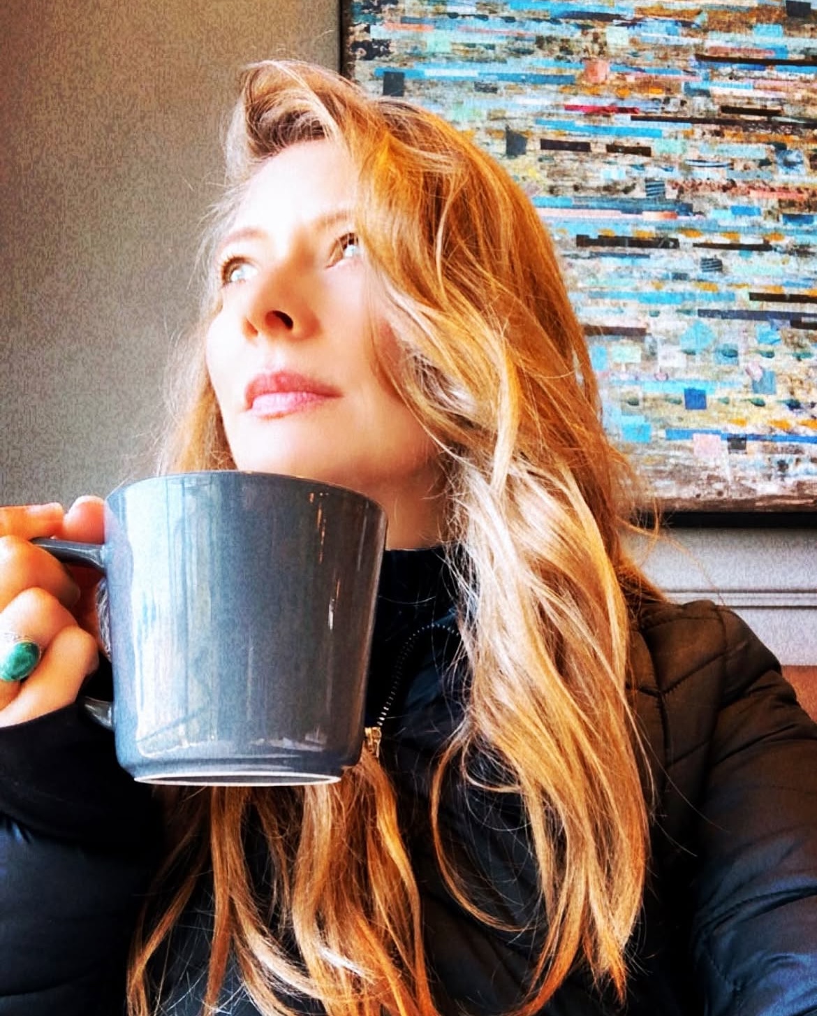 Woman with long wavy hair holding a dark mug, looking to the side, with a colorful textured background.