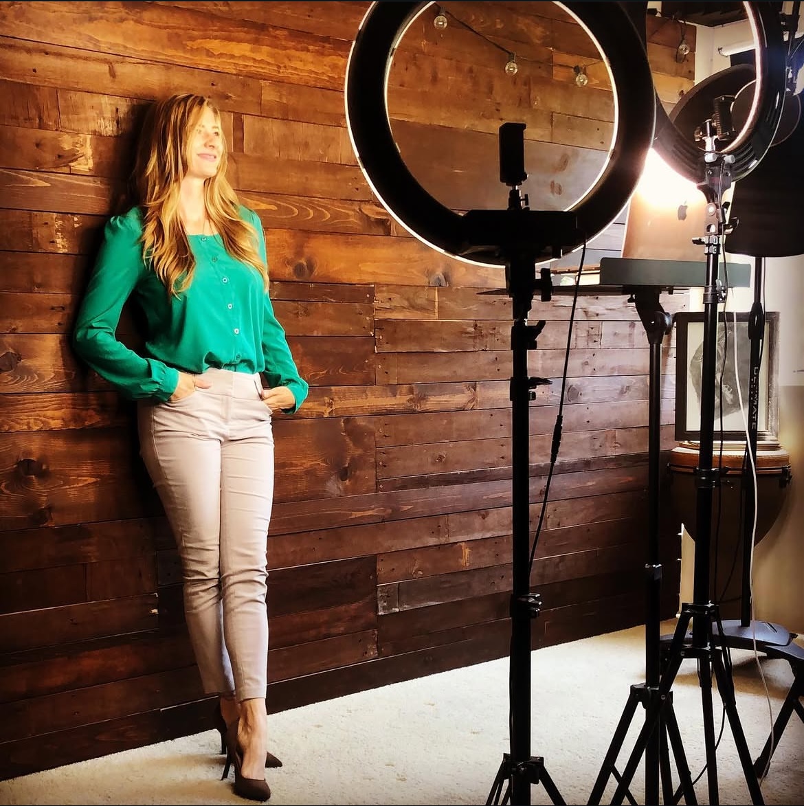 Woman with long hair in a green blouse and beige pants stands against a wooden wall, surrounded by photography equipment.