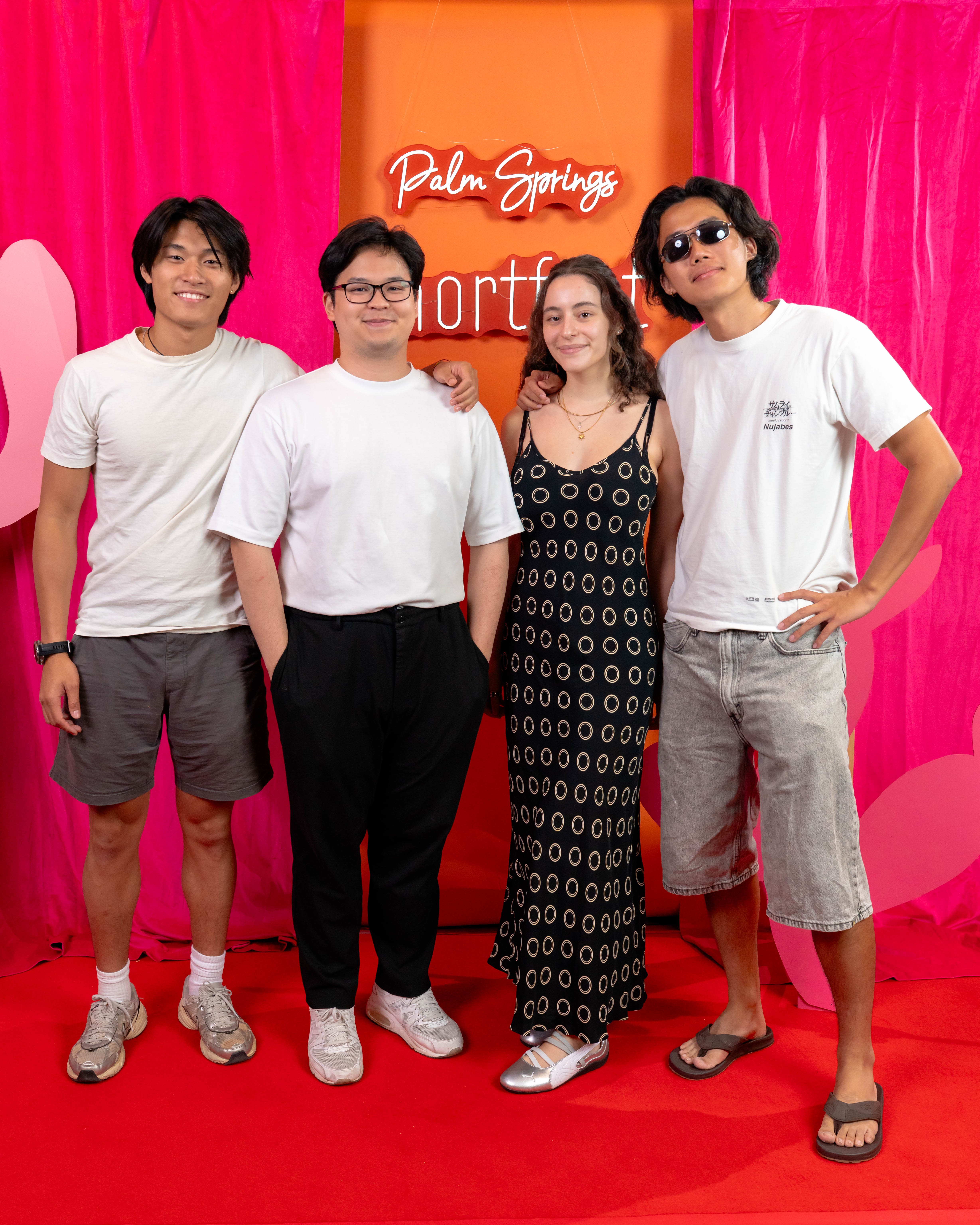 Four people standing on a red carpet in front of pink curtains and an orange backdrop with text, smiling.