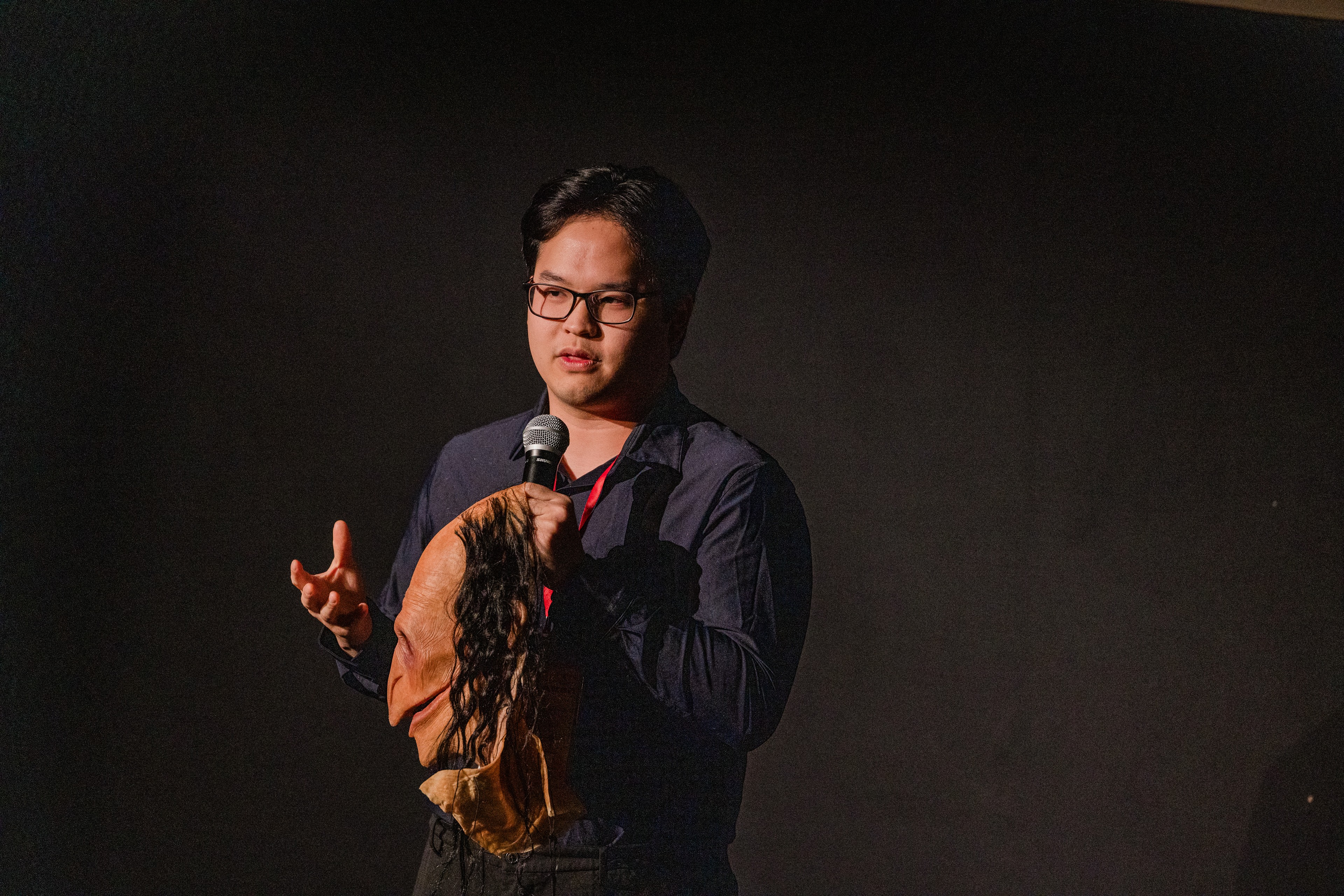 Person speaking into a microphone, wearing glasses and a dark shirt, holding a brown object, on a dark background.