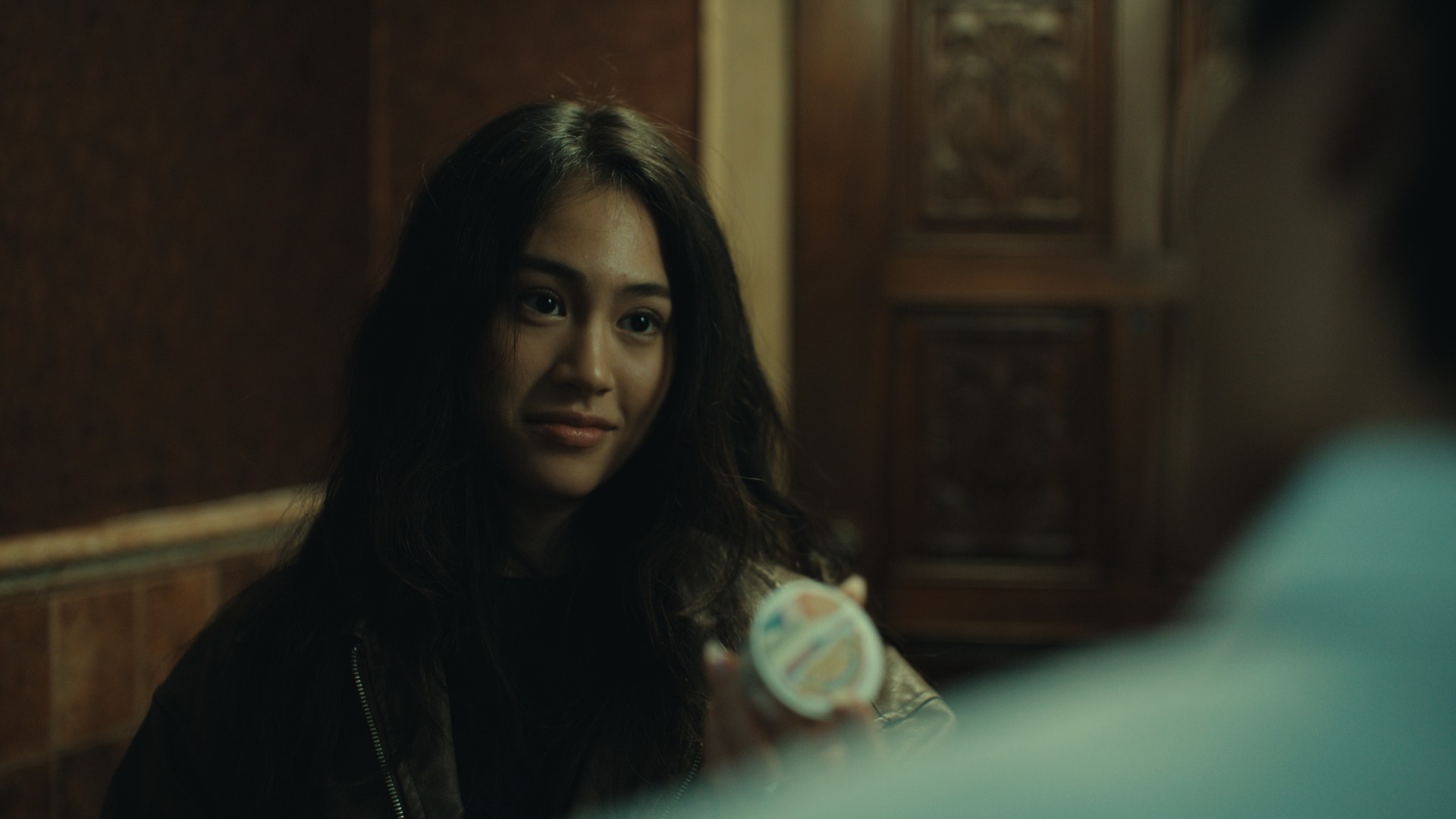 A woman with long dark hair smiling at a person holding a small object, in a dimly lit room.