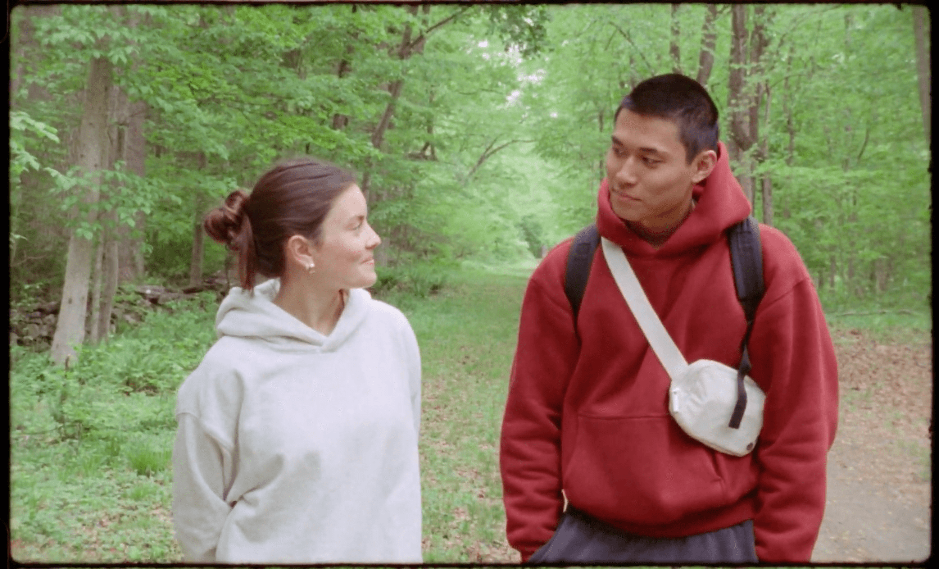 Two people walking and talking in a green forest, one wearing a gray hoodie and the other a red hoodie with a backpack.